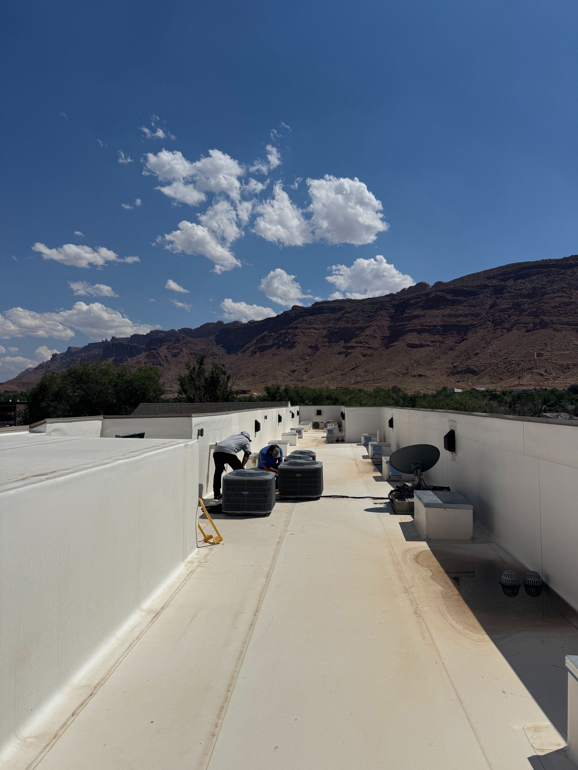 Rooftop view of HVAC units beside a white wall, with mountains and scattered clouds under a blue sky.
