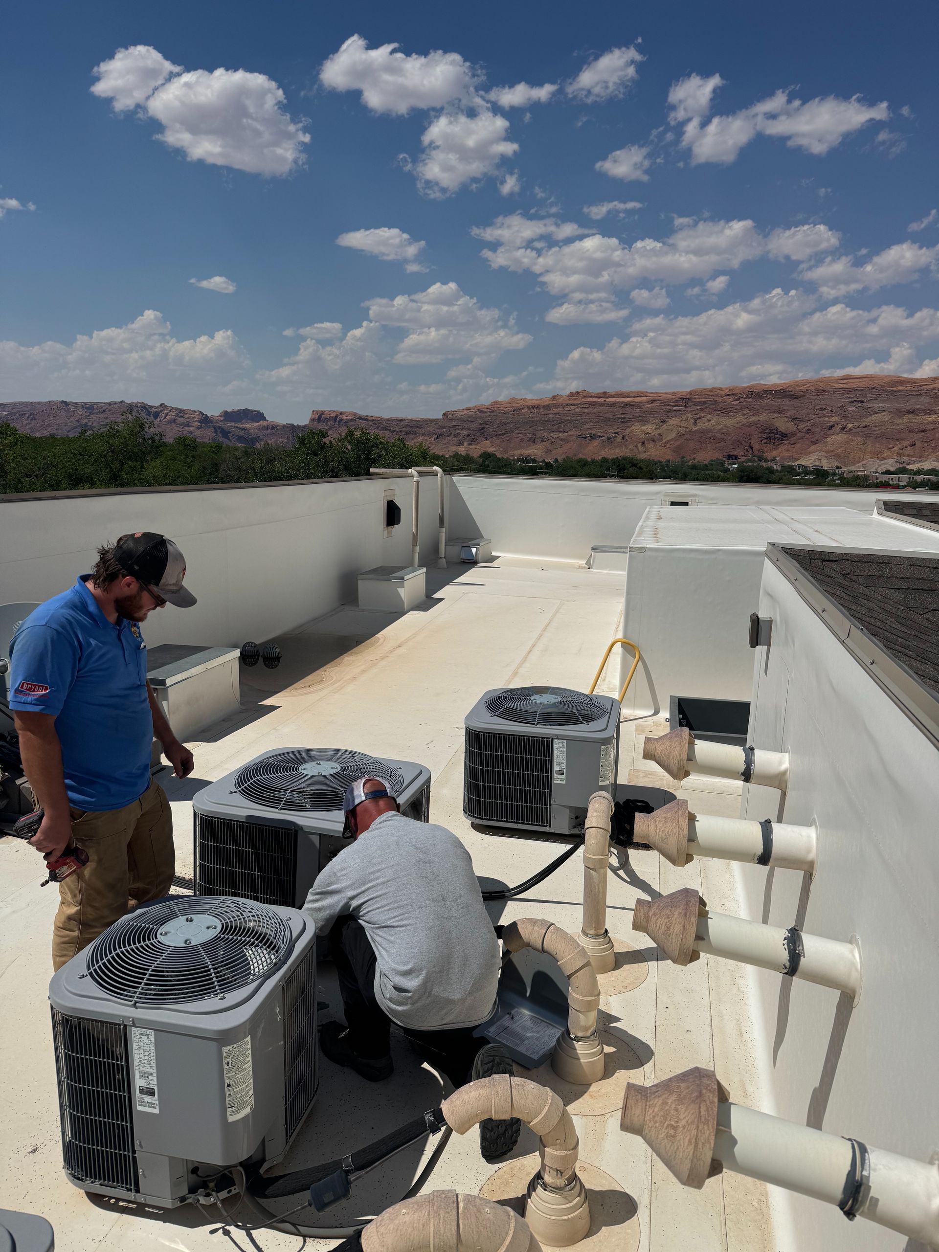 Technicians service rooftop HVAC units on a white building under a sunny sky.