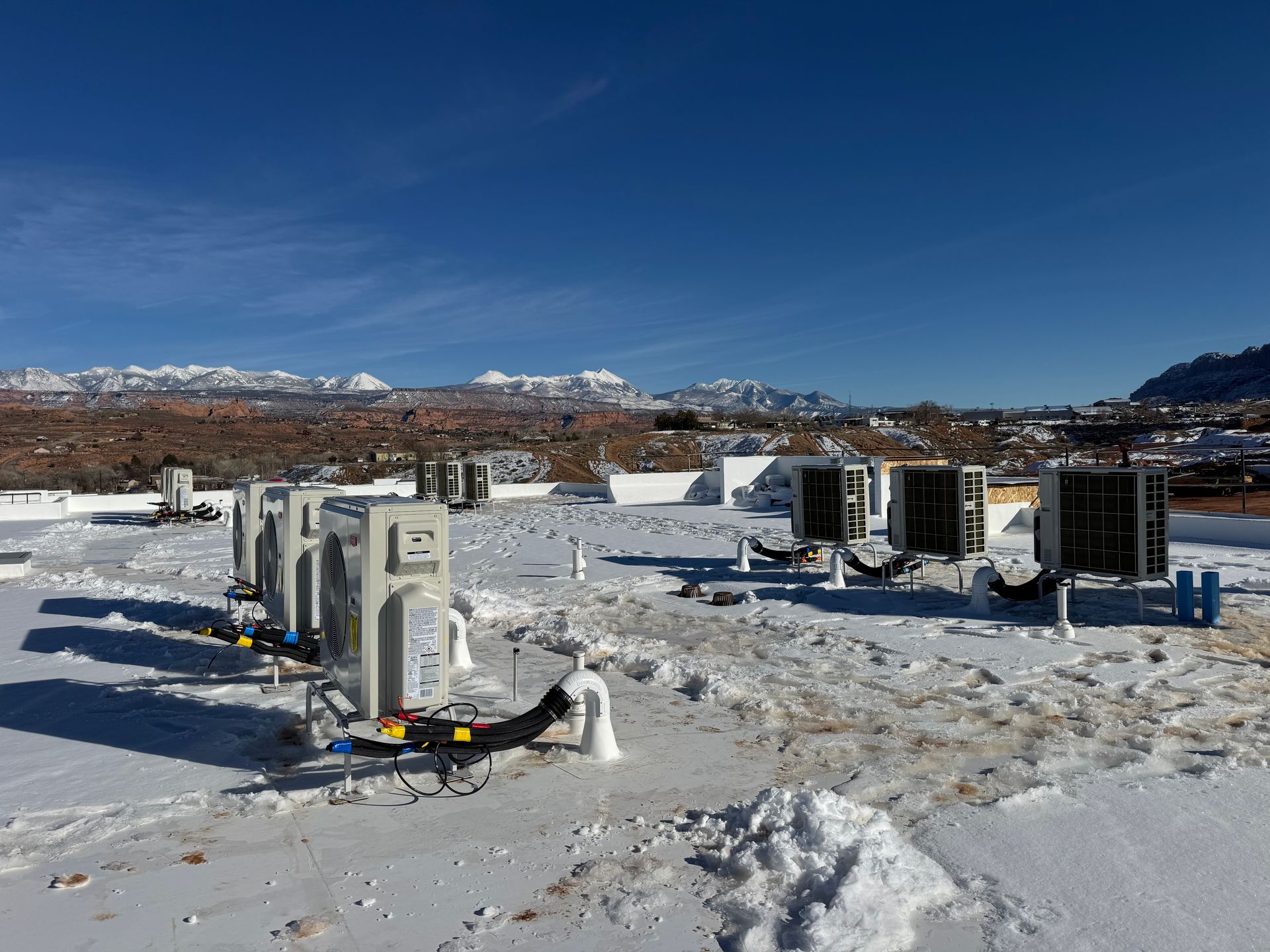 Snowy outdoor utility site with several electrical units under a clear blue sky, mountains in the background