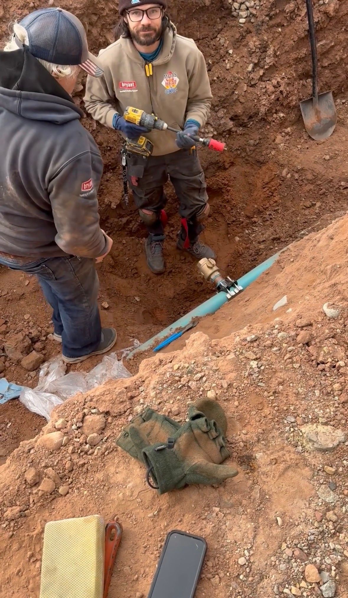 Two workers in a muddy trench examining a blue pipe beside a green bag and tools