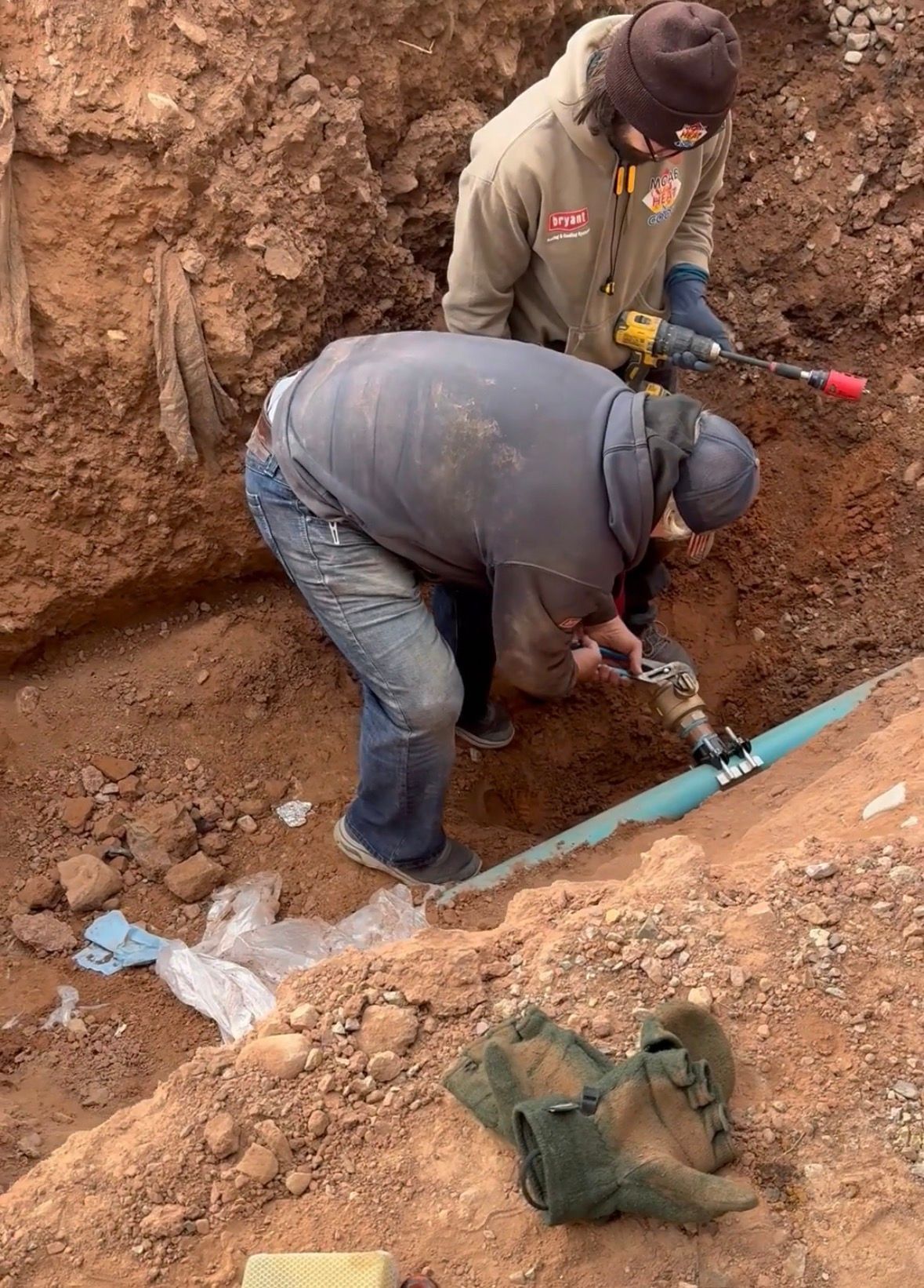 Two workers repairing a blue pipe in a dirt trench, one using a tool while another stands behind.