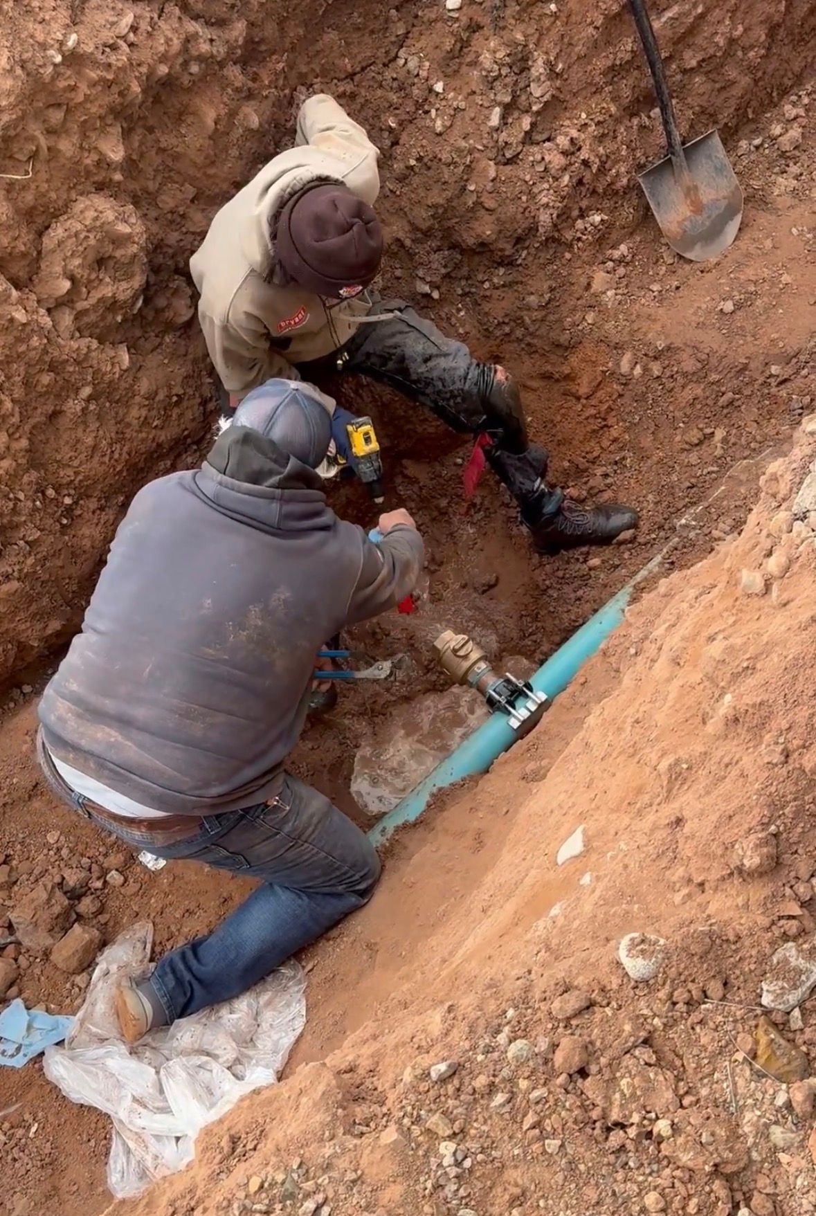 Workers repairing a broken blue pipe in a dirt trench with shovels and tools