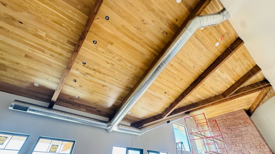 Wooden ceiling with exposed beams and metal ductwork above a partially visible brick wall and windows