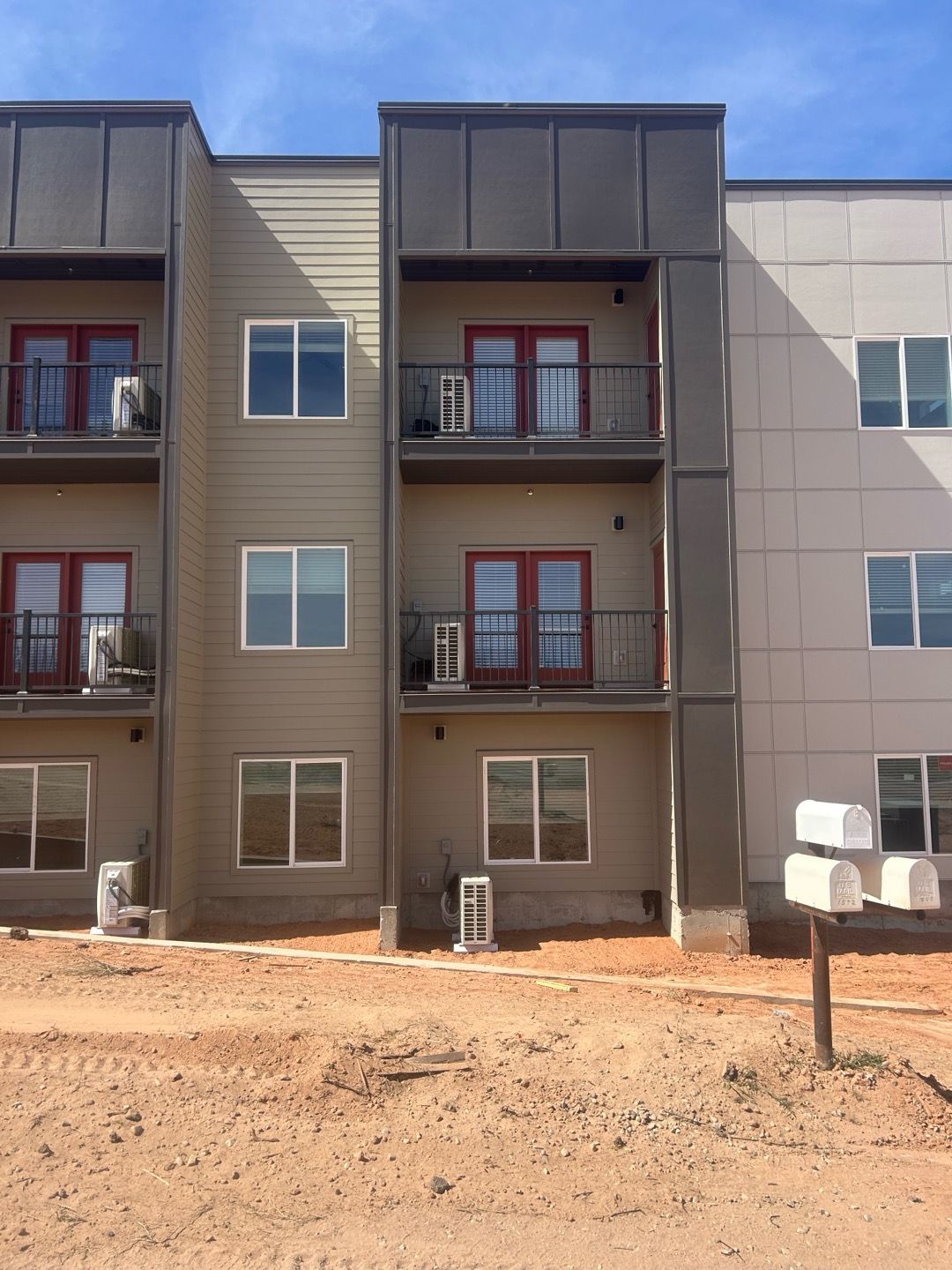 Three-story apartment building with balconies and red railings, viewed from a dirt lot under construction.