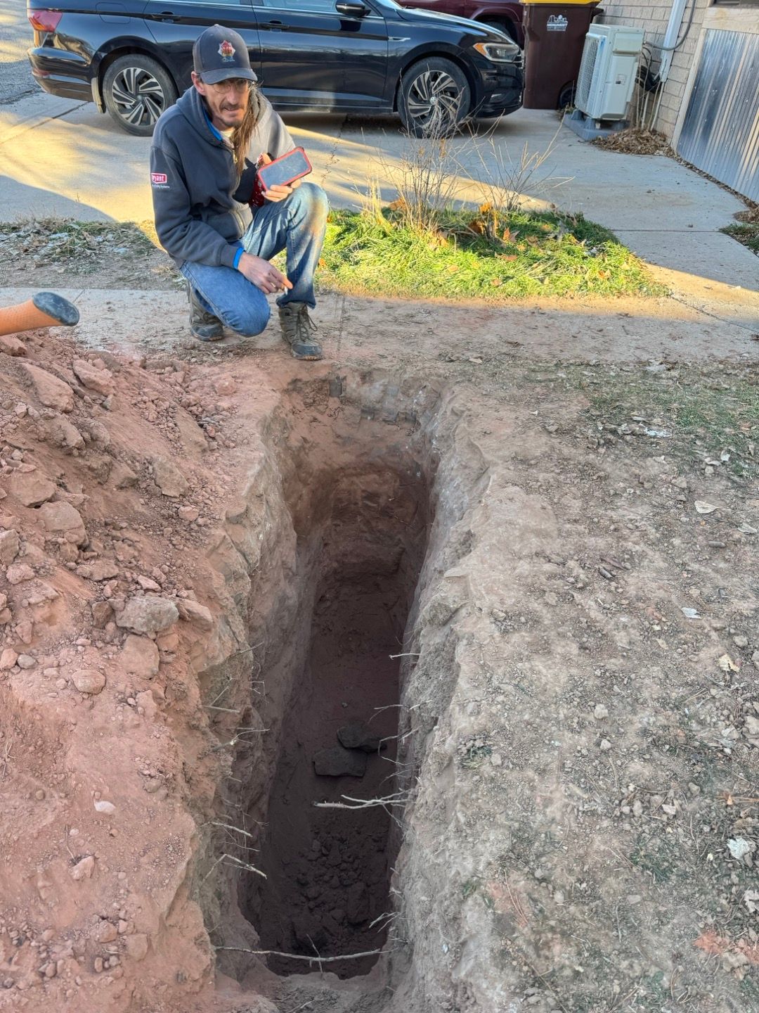 Worker kneeling beside a deep trench cut into a yard near a parked car.
