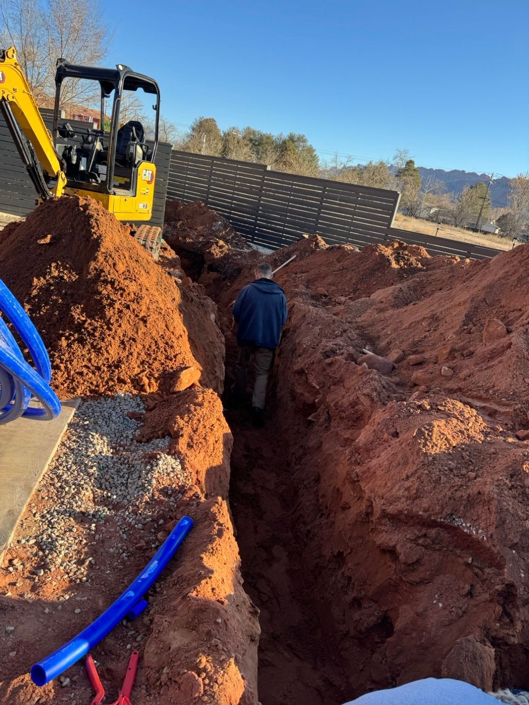 Backhoe and worker in a deep trench at a muddy construction site with red soil and blue pipes