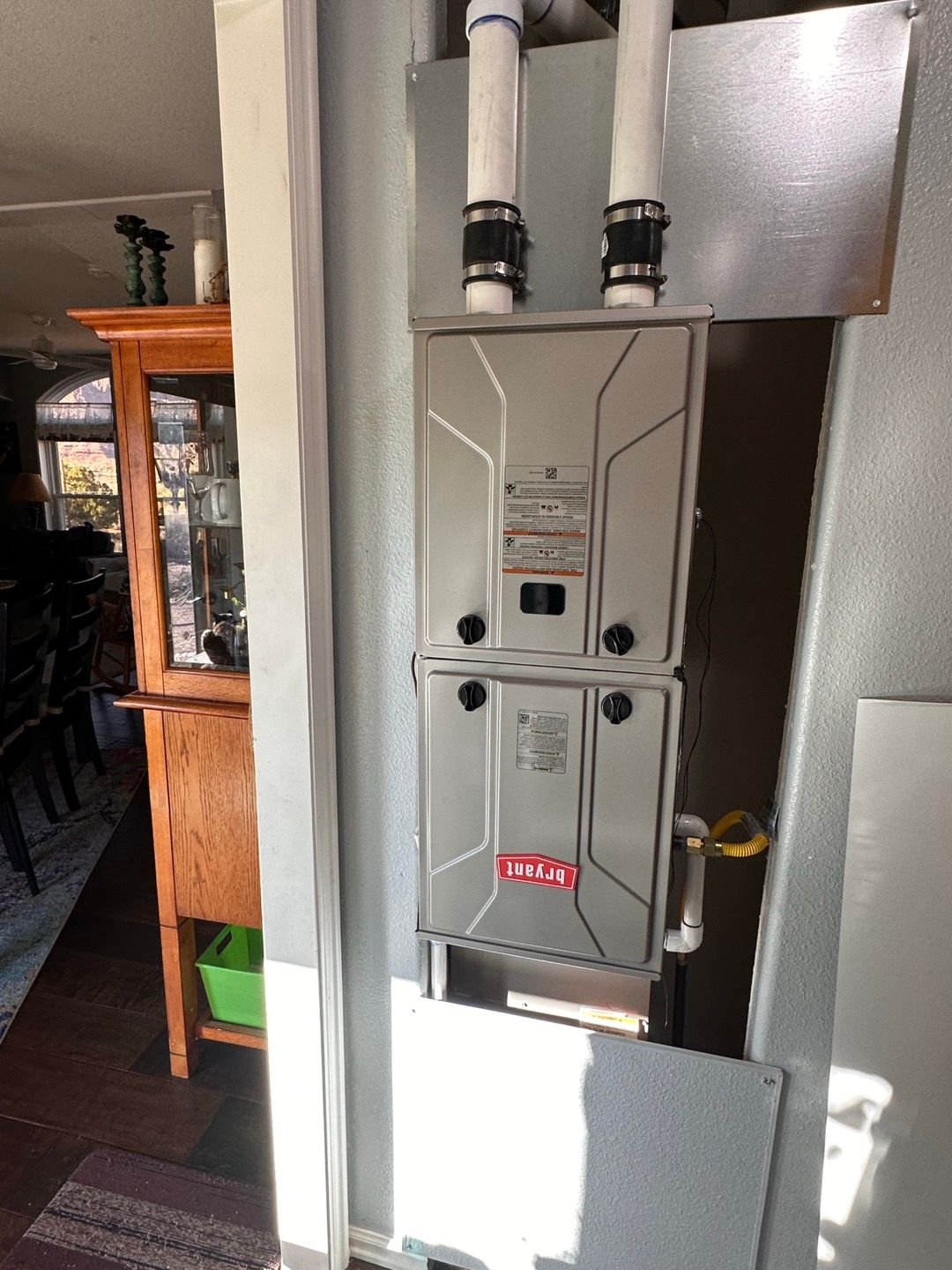 Gray furnace unit with two white vent pipes in a utility closet, beside a wooden cabinet.