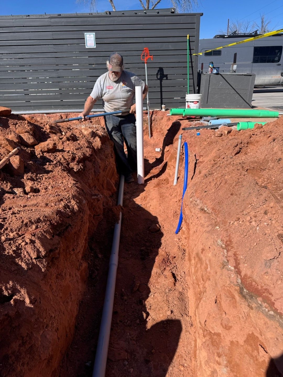 Worker in a trench installing a white pipe beside red dirt and a gray house.