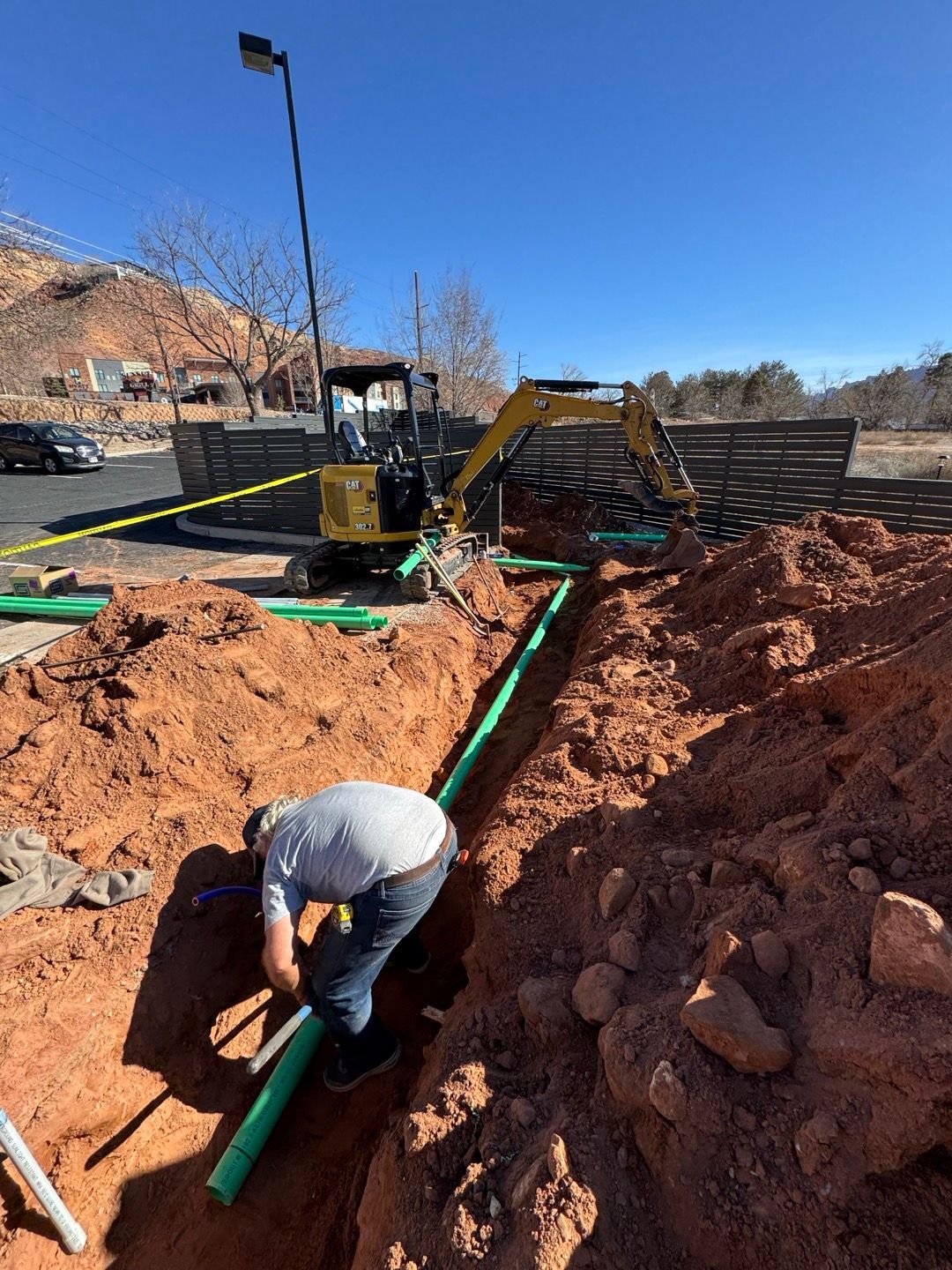 Worker in a trench installing green pipes at a construction site under a clear blue sky.