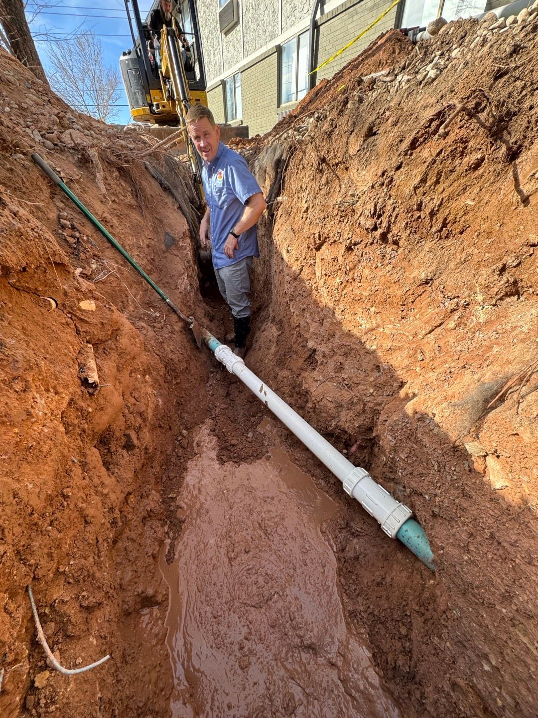 Worker standing in a deep trench beside a white pipe near a building