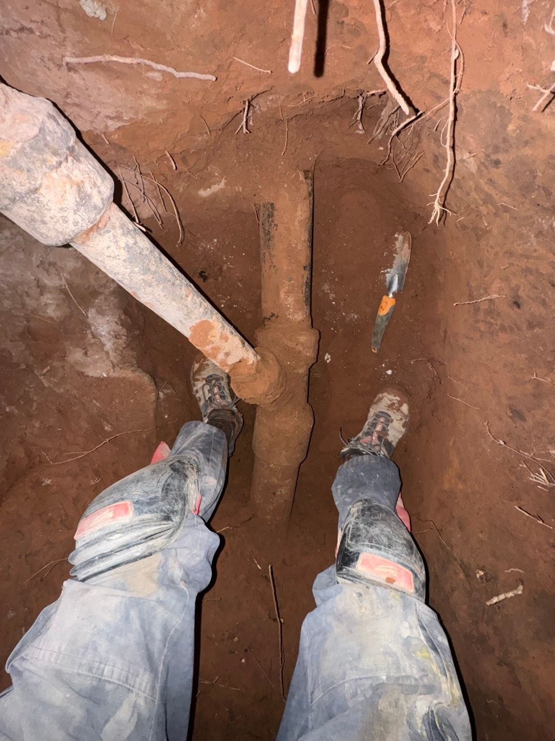 Person standing in a narrow dirt trench with digging tools and boots visible.