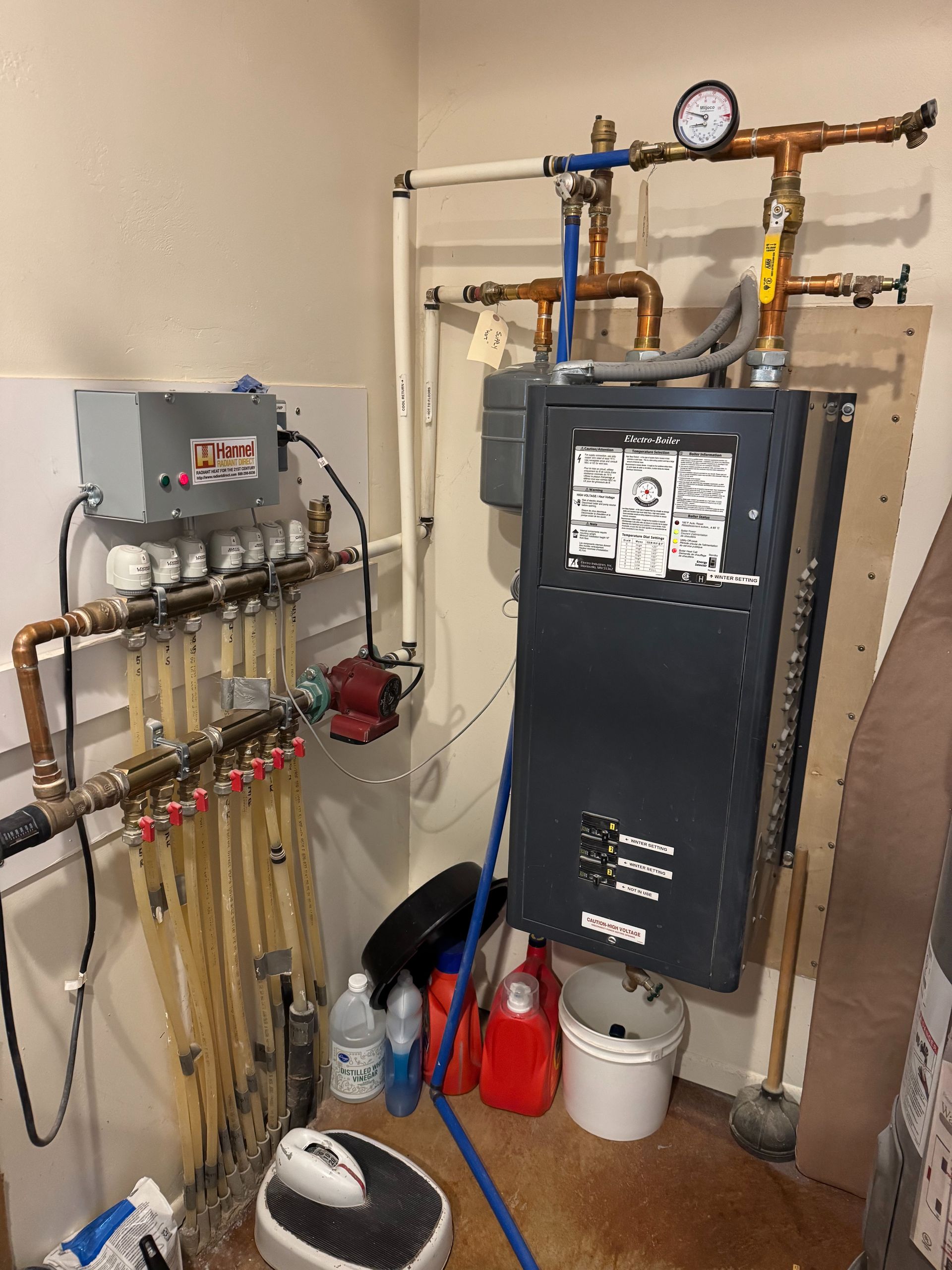 Utility room with boiler, water heater, pipes, gauge, and cleaning supplies on the floor.