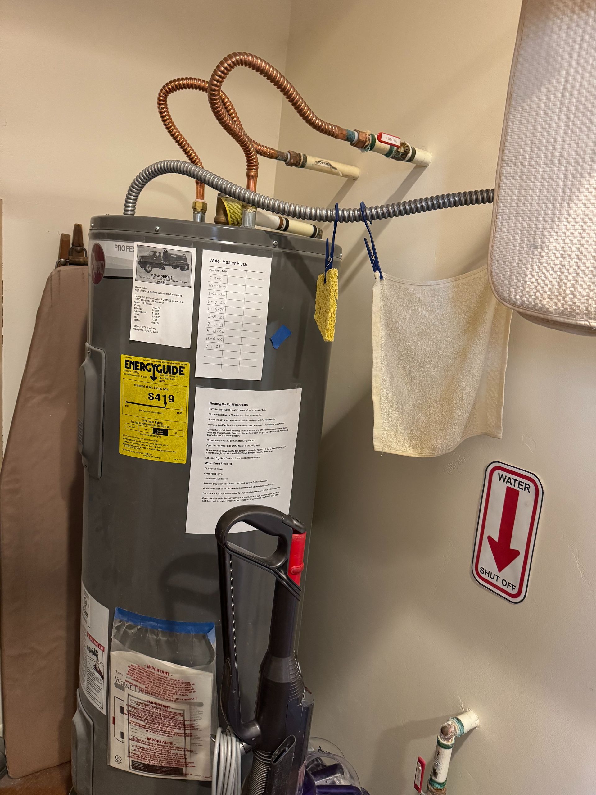 Water heater with attached pipes and labels beside a vacuum cleaner in a small utility area.