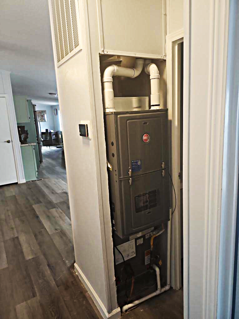 Open HVAC furnace in a narrow hallway closet with exposed pipes and vents
