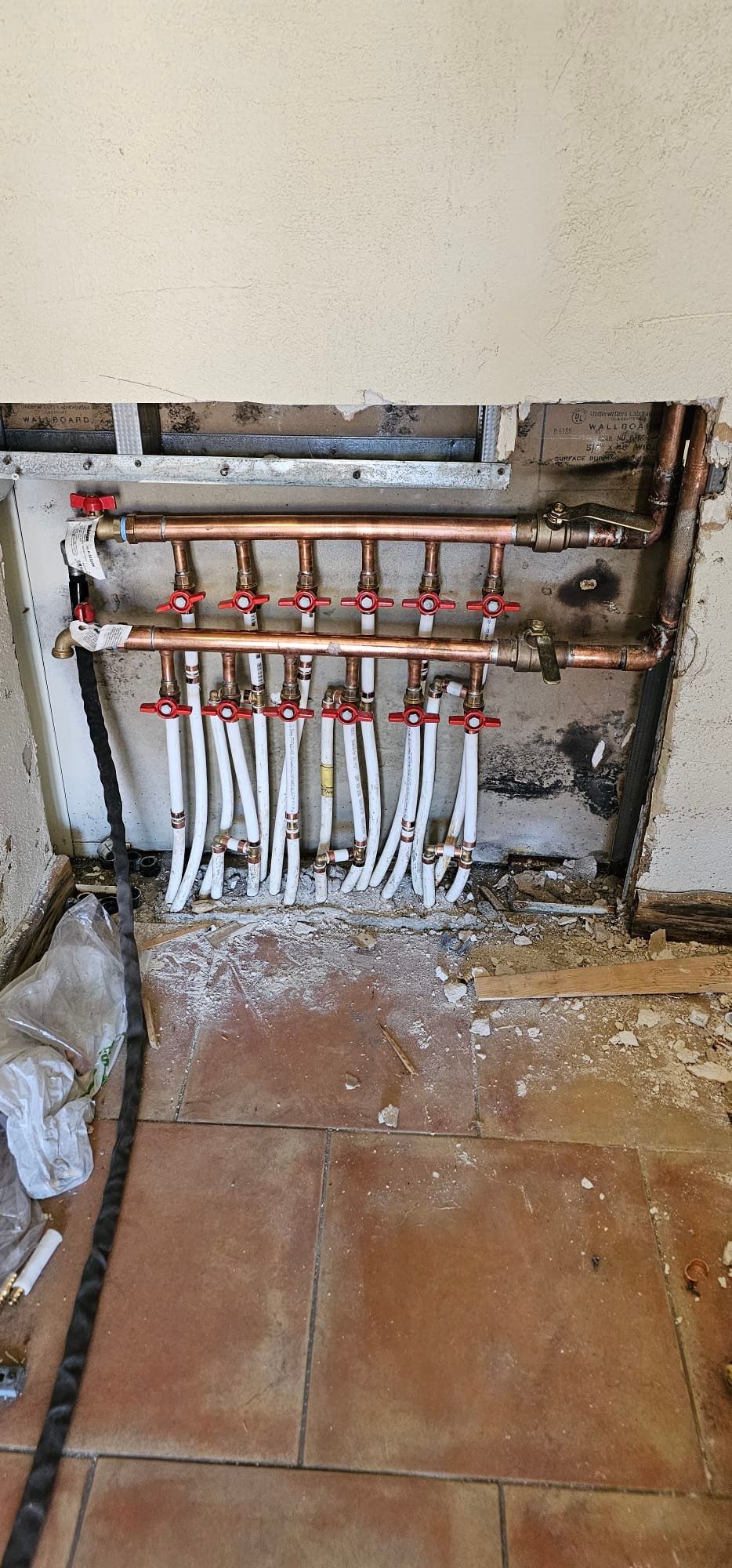Exposed plumbing manifold with multiple red valves and white pipes in a wall recess above a tiled floor