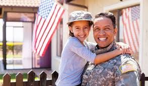 Smiling soldier in camouflage uniform giving a piggyback ride to a child outdoors with American flags