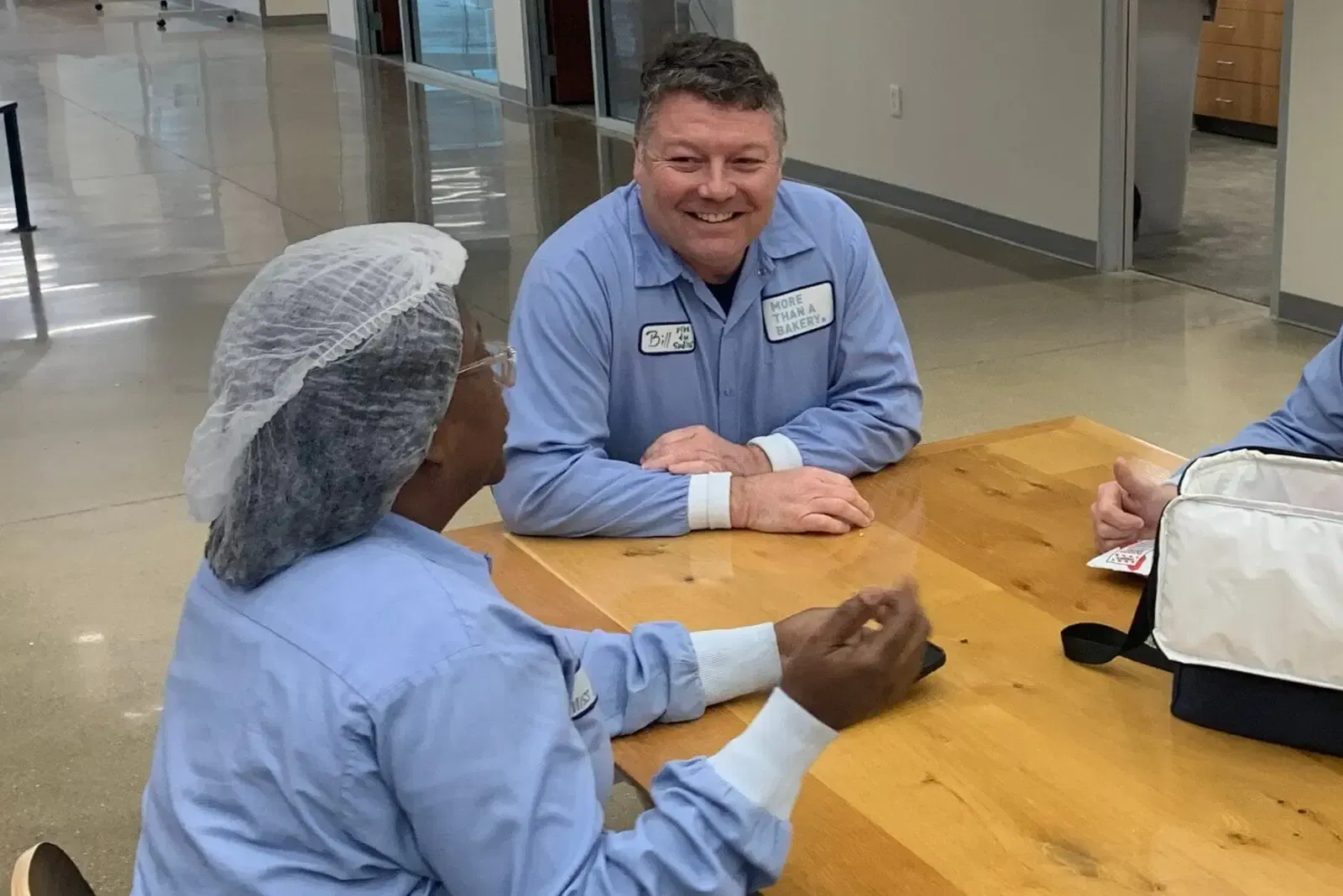 Two people in blue uniforms at a table. One smiles; the other is wearing a hairnet. In a cafeteria.