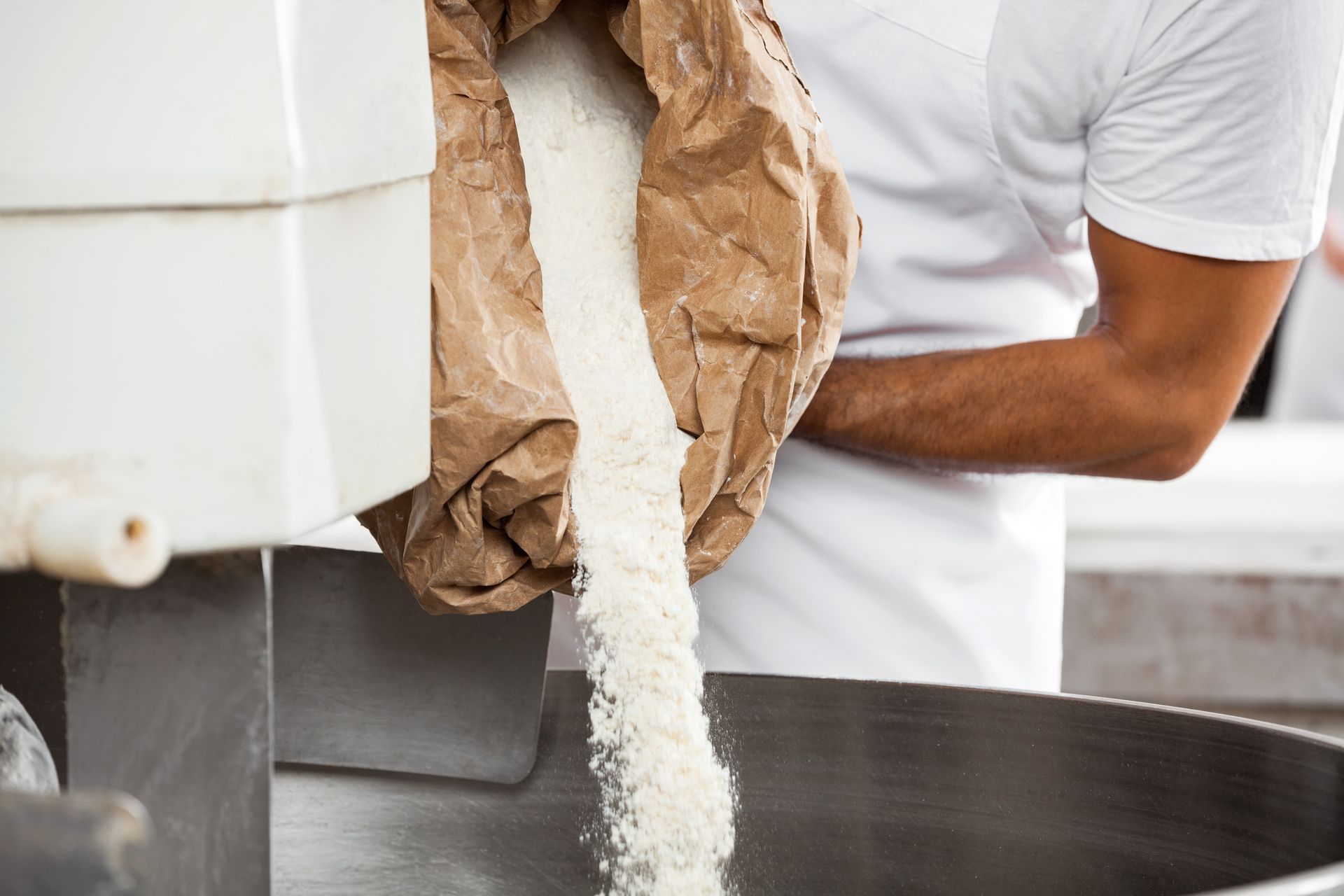 Baker pouring flour from a brown paper bag into a large stainless steel bowl.