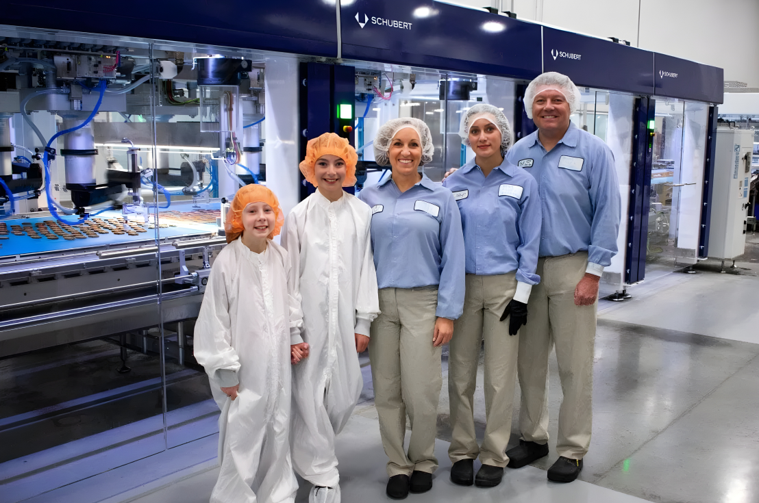 Three people in blue shirts and 2 people in white bunny suits smiling and standing in front of a robotic packaging system.