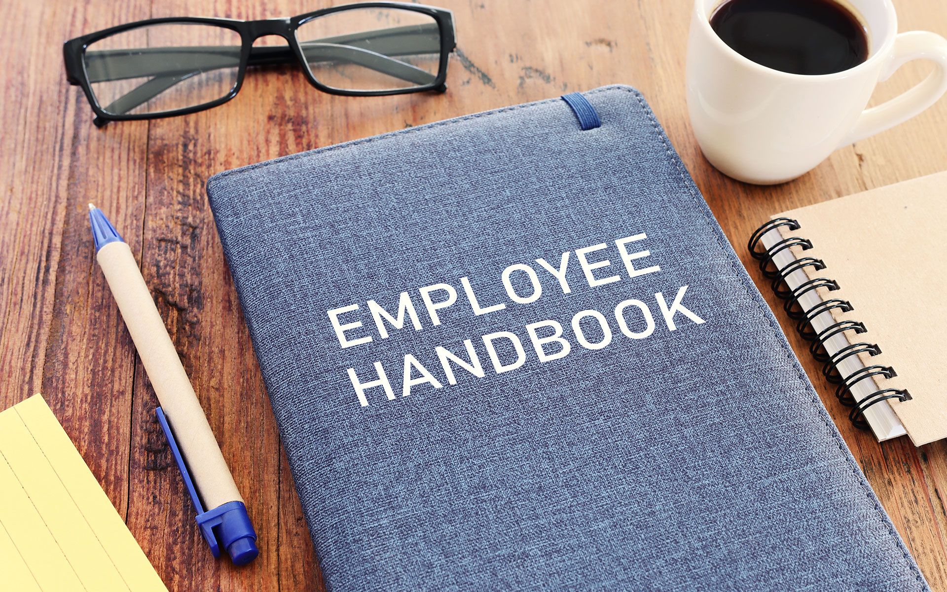 An employee handbook is sitting on a wooden table next to a cup of coffee and glasses.