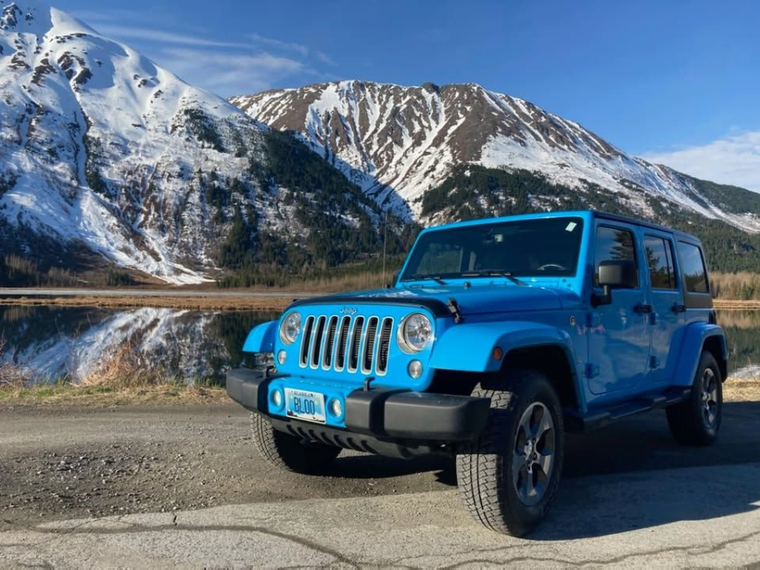 Bright blue Jeep Wrangler parked on a road with snowy mountains and a lake in the background
