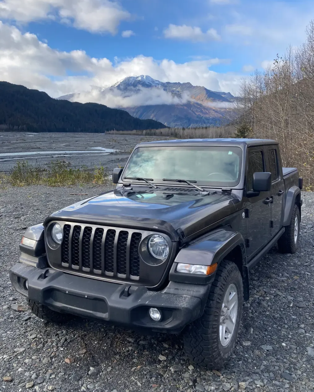 Black Jeep Wrangler parked on a rocky riverbank with mountains and clouds in the background