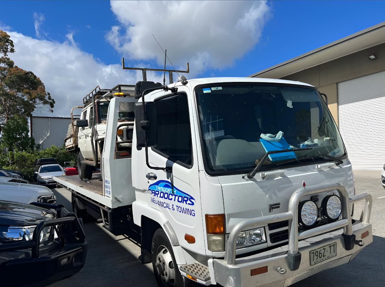 A White Tow Truck With a Flatbed is Parked in Front of a Building — Skazas Automotive In Cardiff, NSW