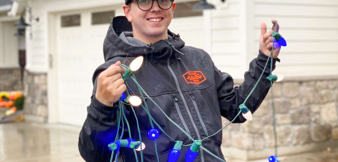 Person holding a tangled string of colorful holiday lights outdoors, smiling near a house