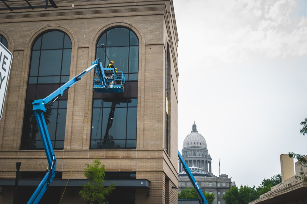 Workers in blue lifts cleaning a building facade, with a domed capitol in the background.