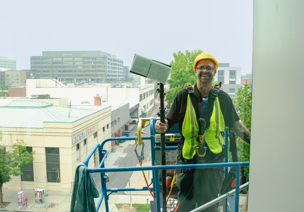 Construction worker on a lift at a building exterior, wearing a hard hat and safety vest.