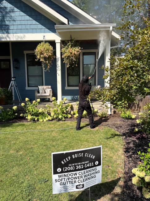 Person trimming shrubs in front of a blue house with a porch and hanging baskets