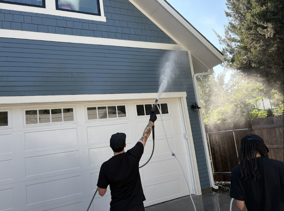 Two people pressure-wash a blue garage exterior in bright sunlight