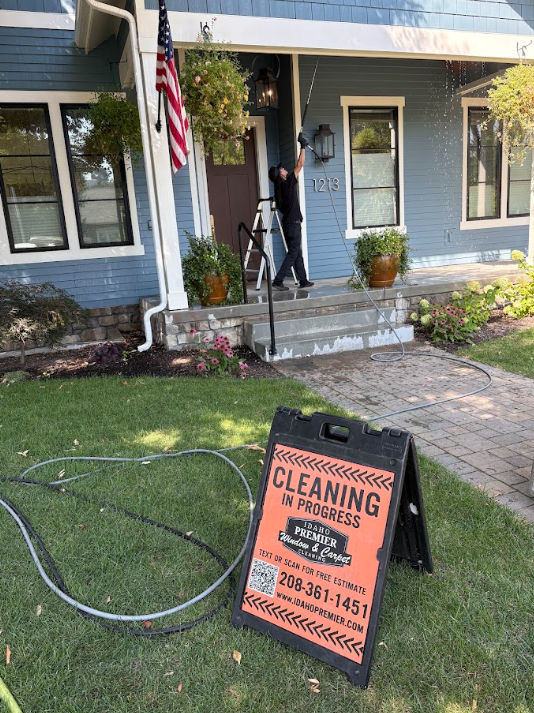Worker power-washing blue house porch; orange cleaning sign on lawn and American flag by the front door.