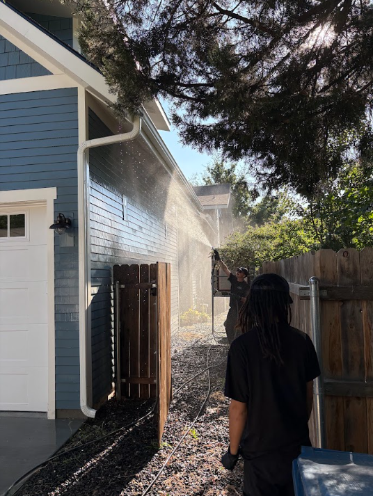 Person walking beside a blue house as water sprays from the roof under a tree.