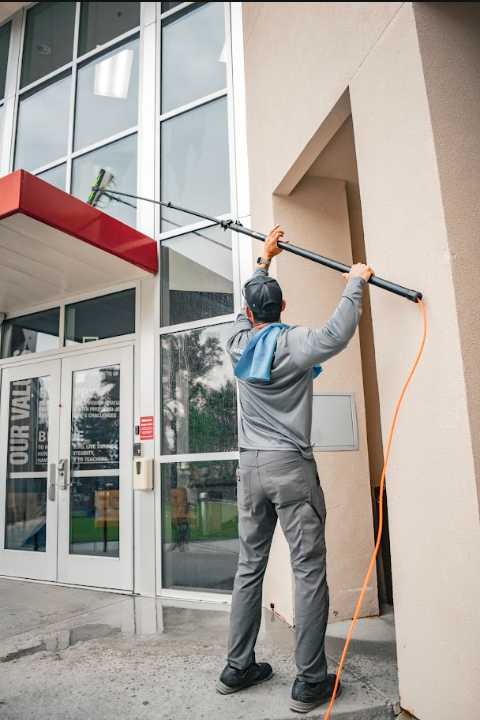 Worker washing windows on a modern building exterior with a long pole and orange hose