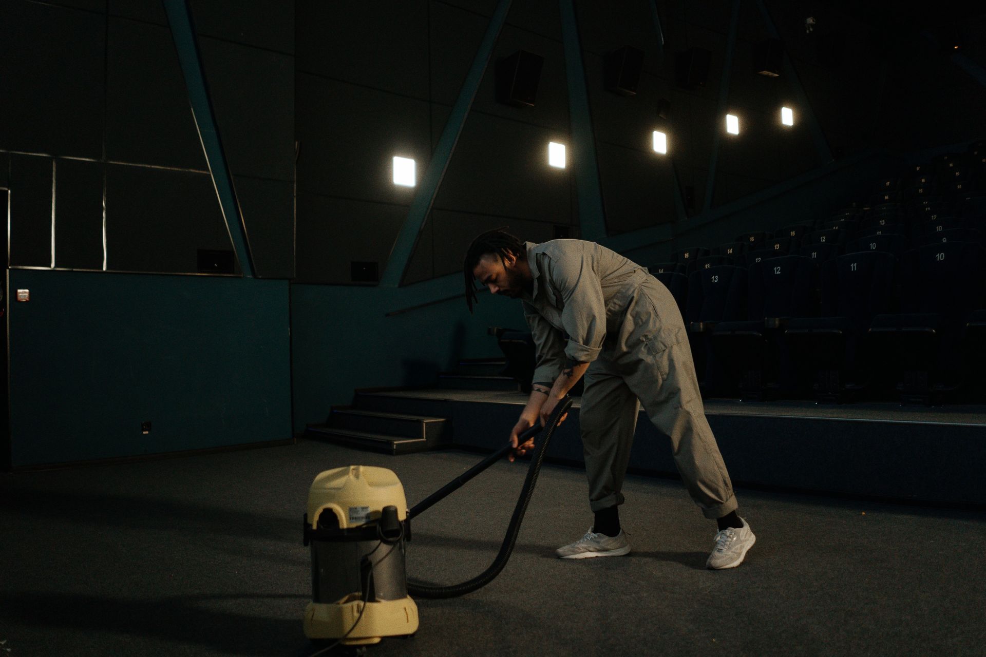 Person vacuuming a dark indoor stadium floor beside a yellow canister vacuum