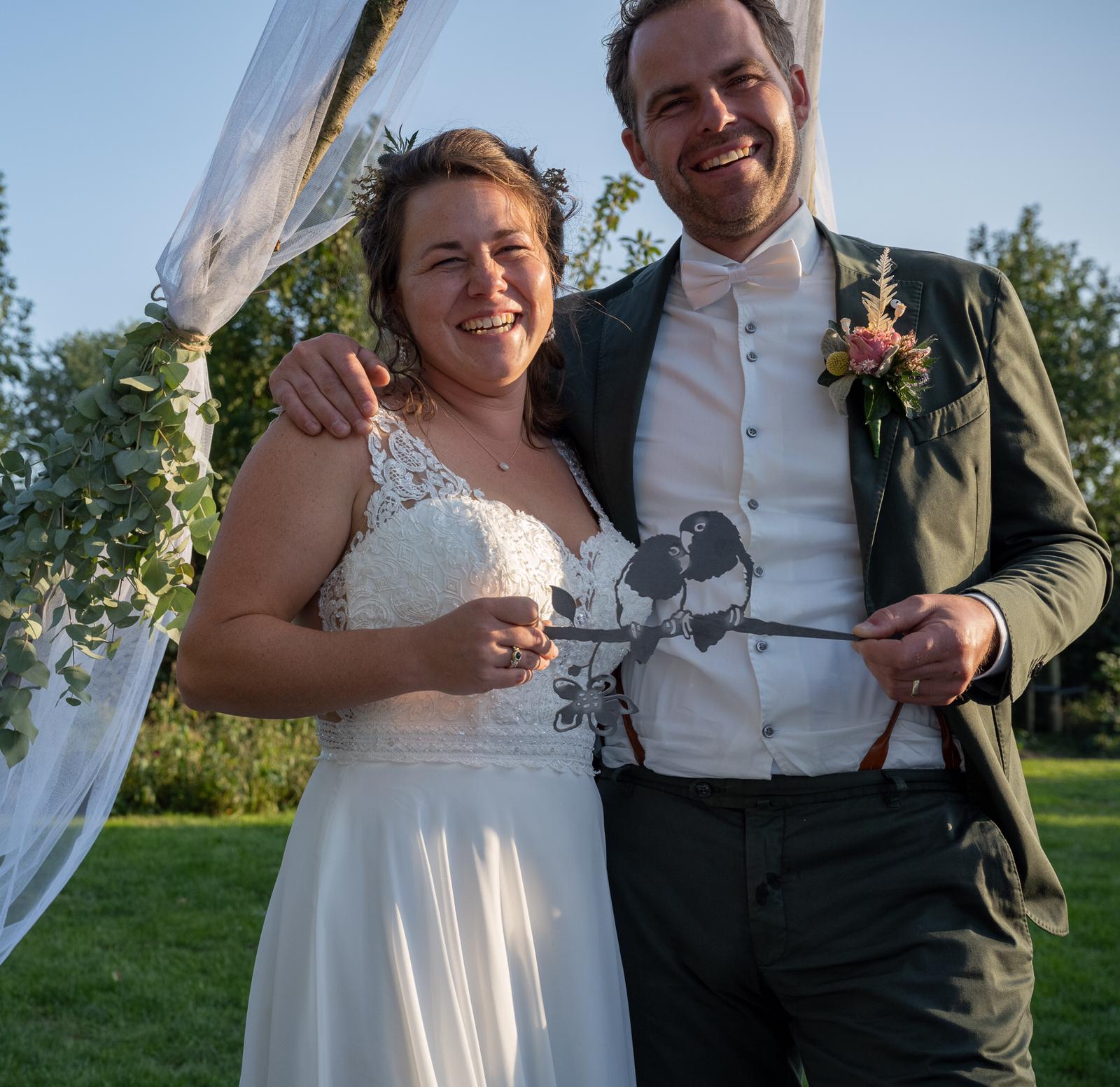 Glimlachend bruidspaar met vogelversiering in de hand; ceremonie in de buitenlucht.