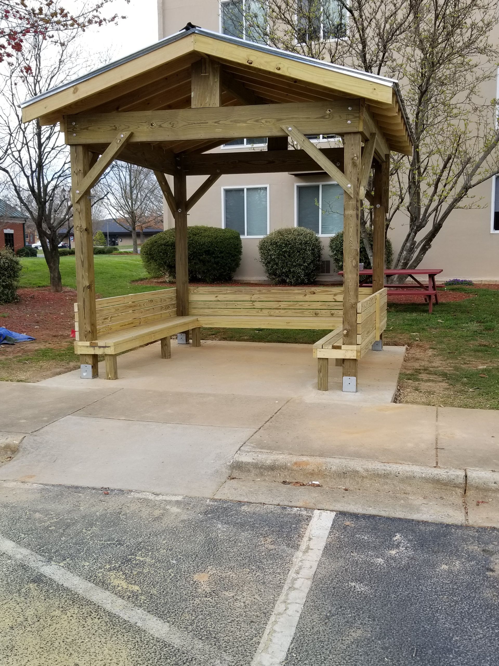 Wooden gazebo with built-in benches, on a concrete pad, in front of an apartment building.