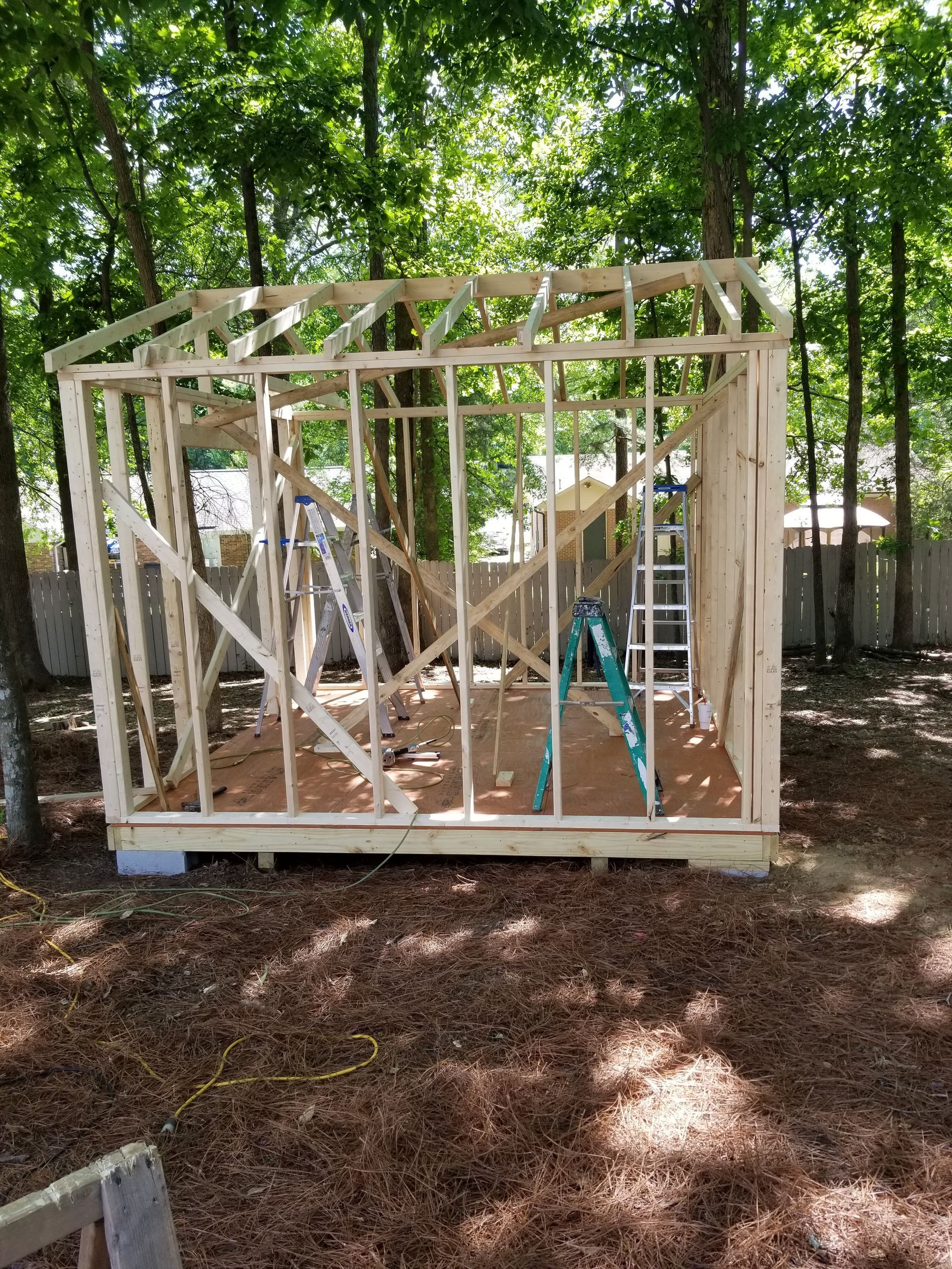 Framing of a wooden shed under construction, set in a wooded backyard with a ladder.