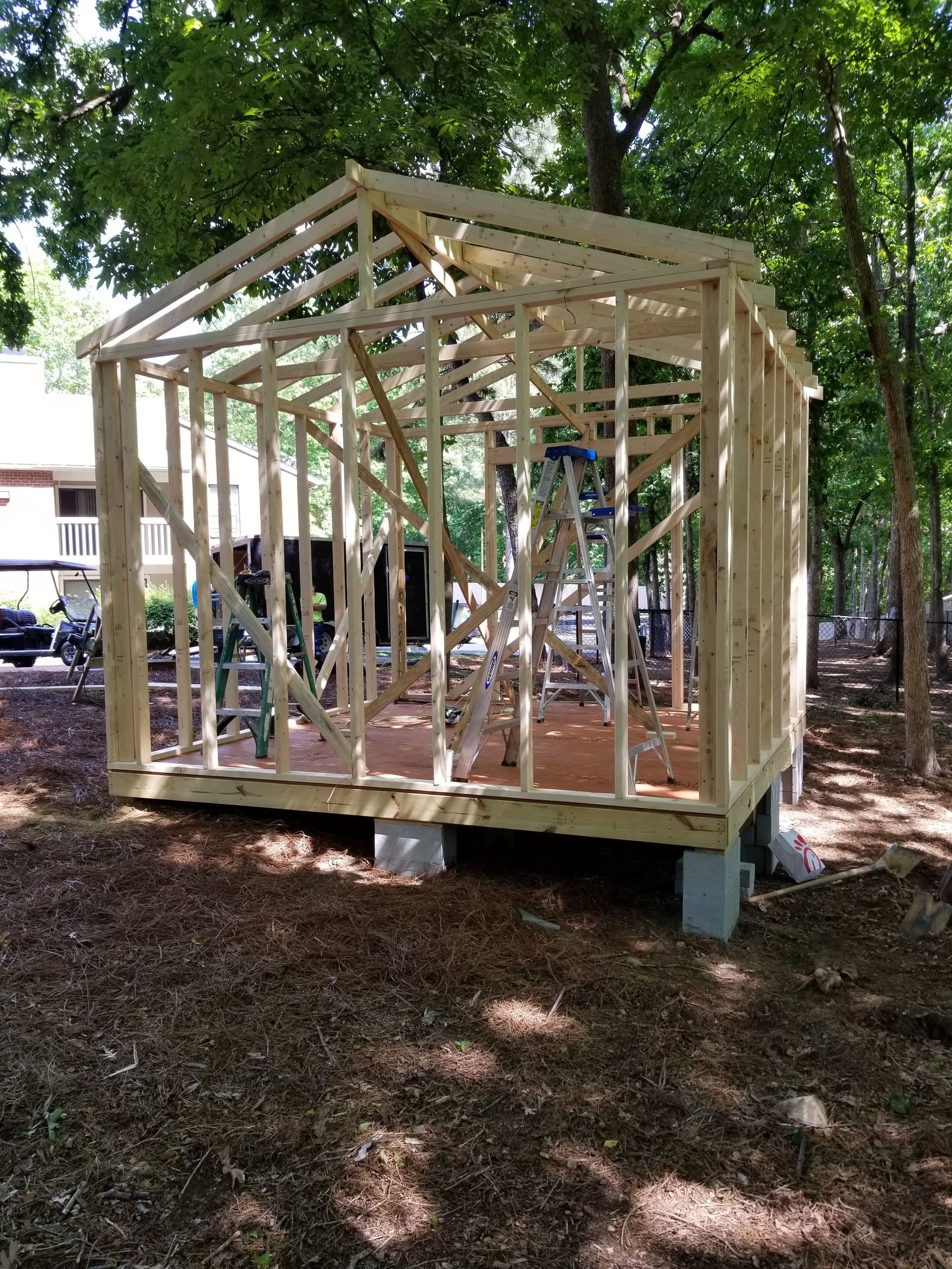 Wooden shed frame under construction, sitting on concrete blocks, outdoors in a wooded area.