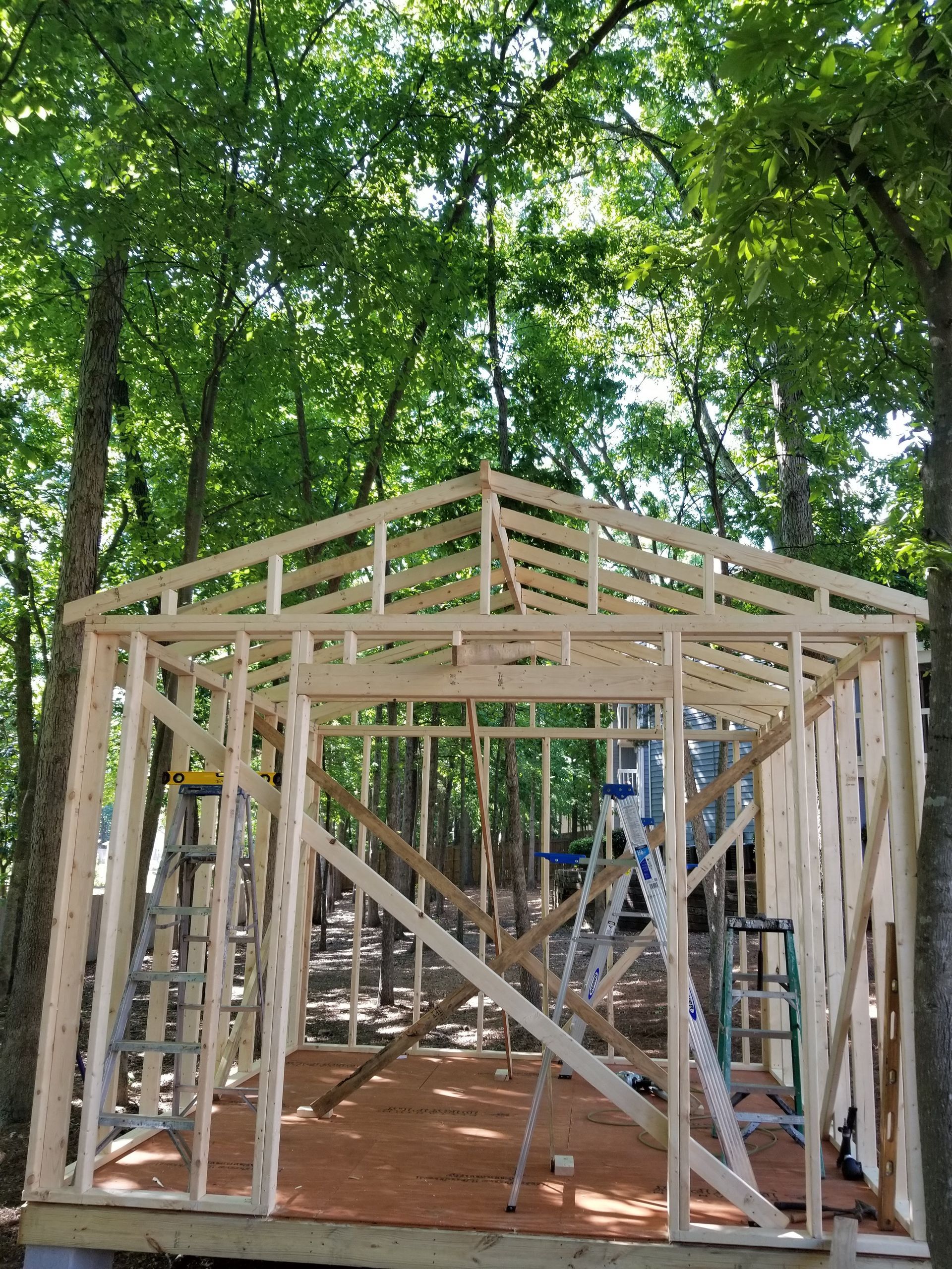 Wooden shed frame under construction in a wooded area; ladder and tools visible.