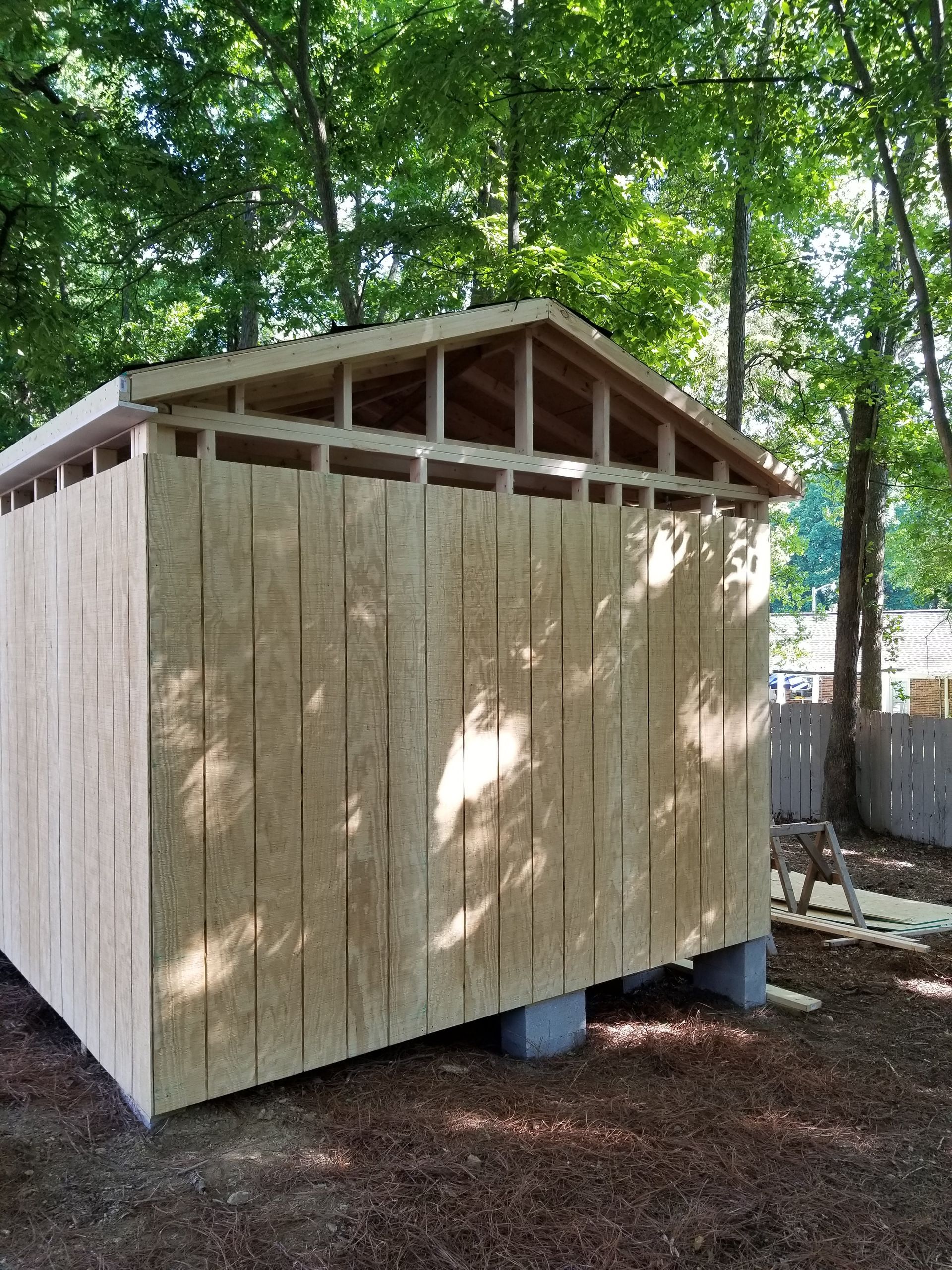 Wooden shed under construction, light brown siding, unfinished roof, surrounded by trees.