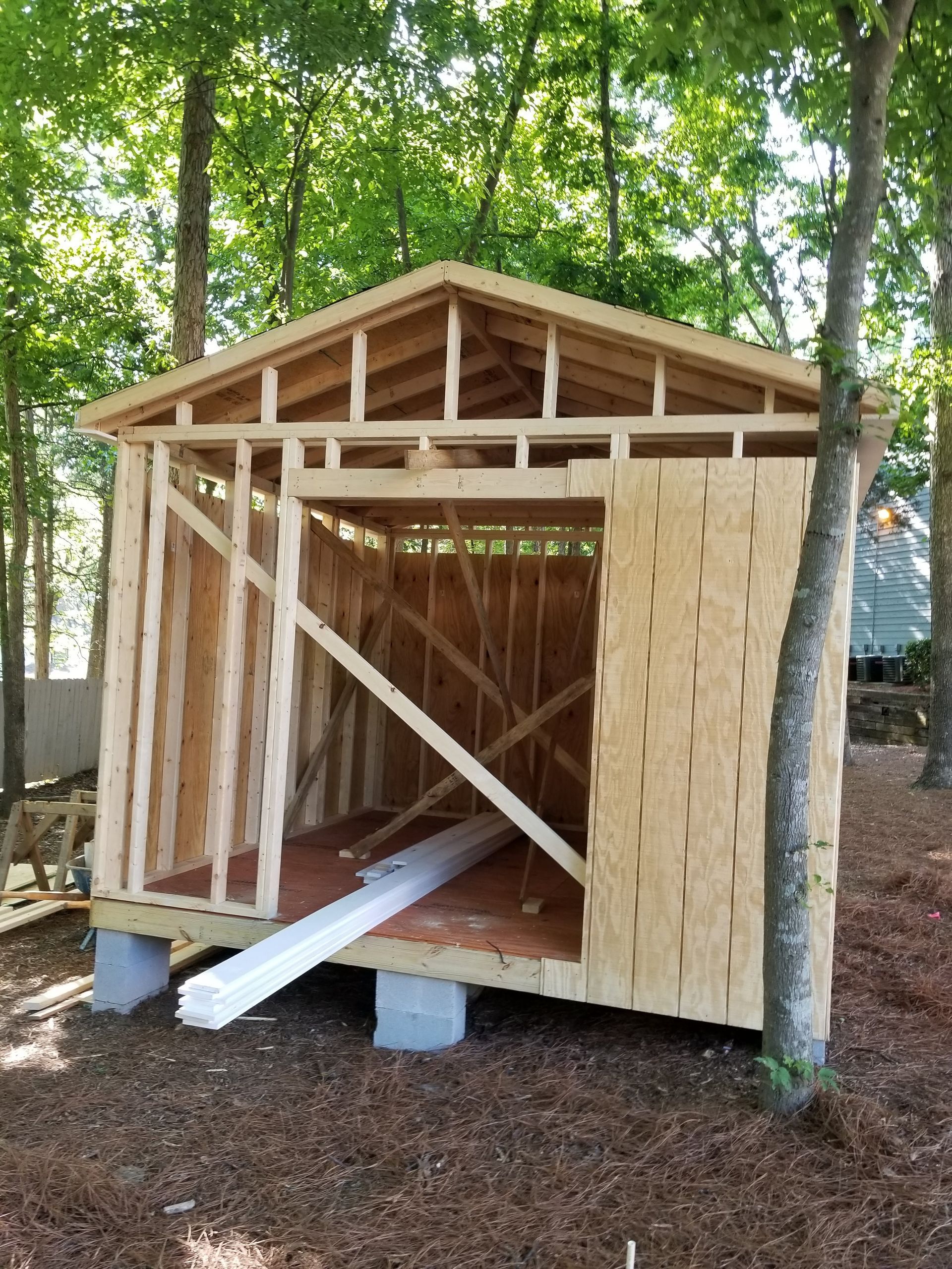 Wooden shed under construction in a wooded area, with partially installed wall and roof framing.