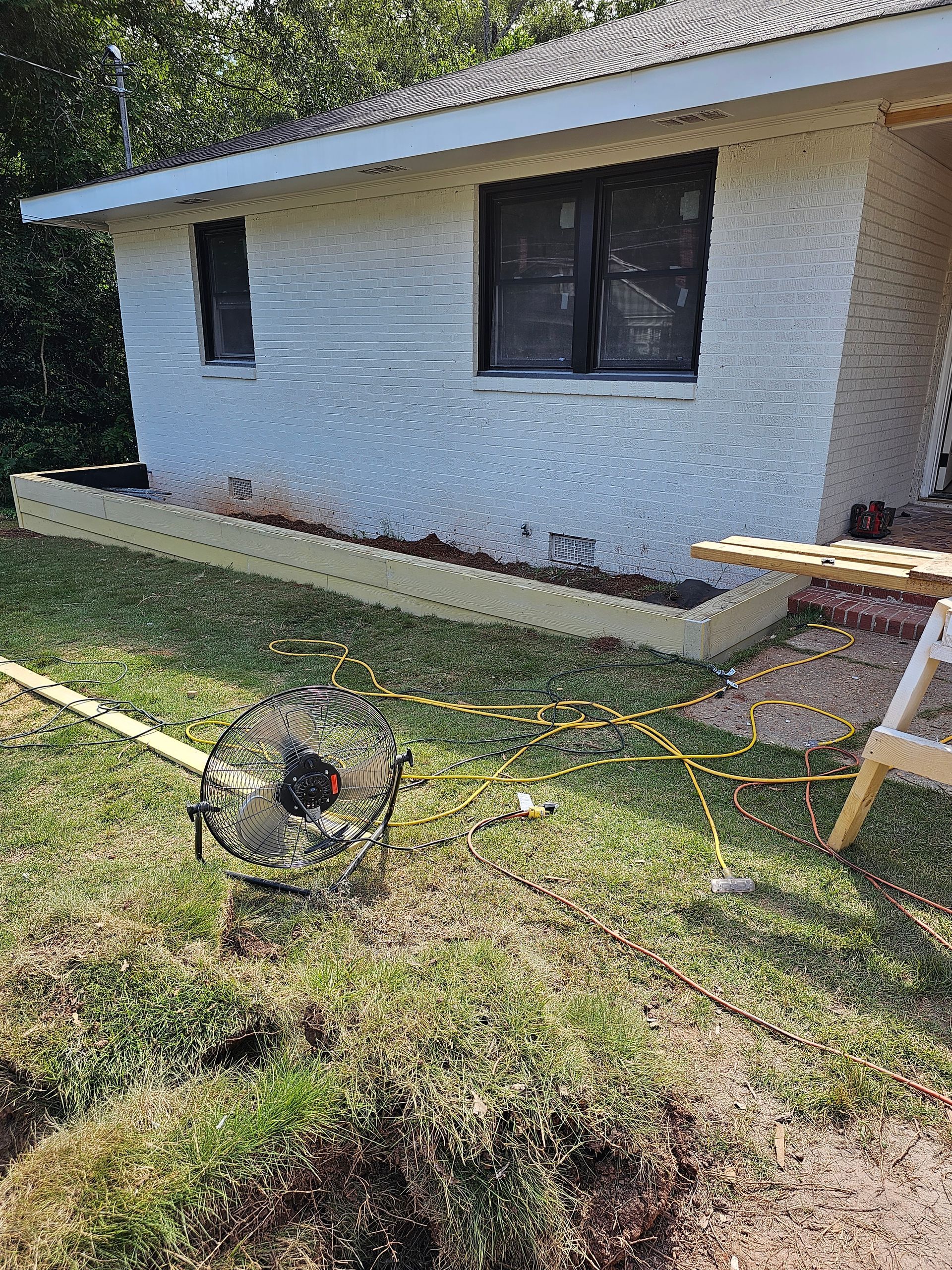 A circular saw on grass in front of a white building with black window frames, wooden board below.