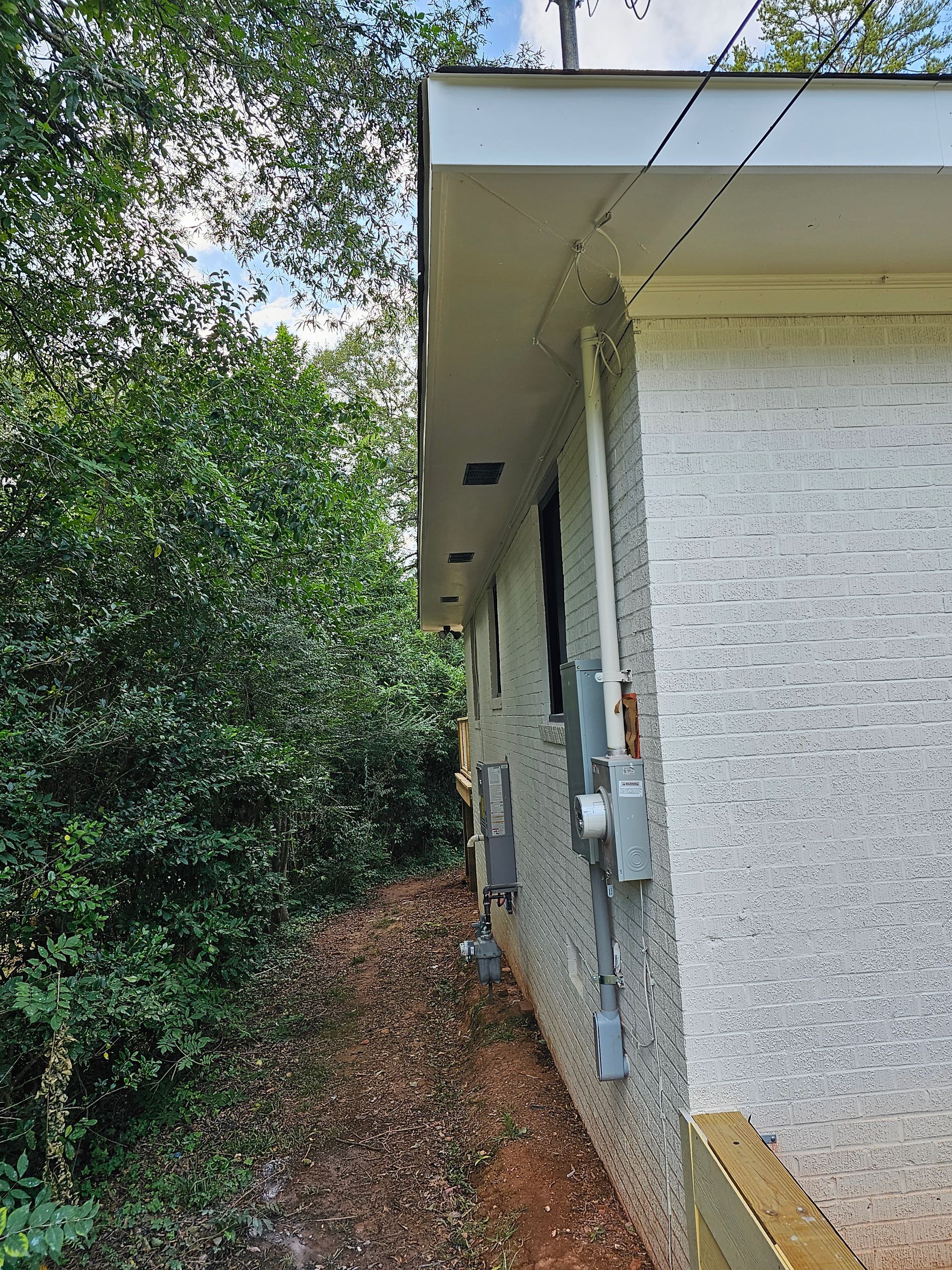 Side view of a white stucco house with electrical boxes. Green trees and sky are visible.