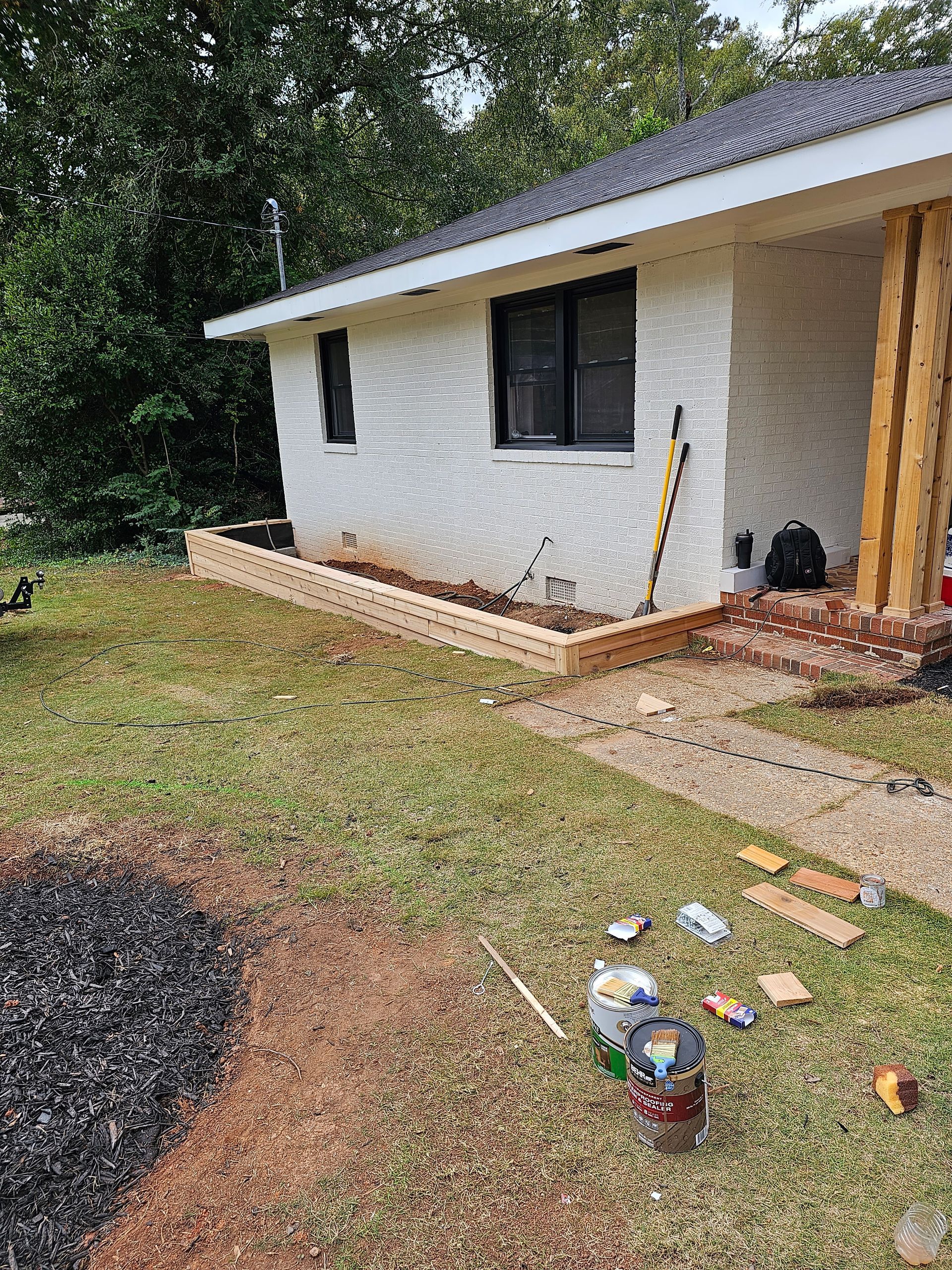 A white house with a brick planter in front, a pathway, and construction supplies scattered on the grass.