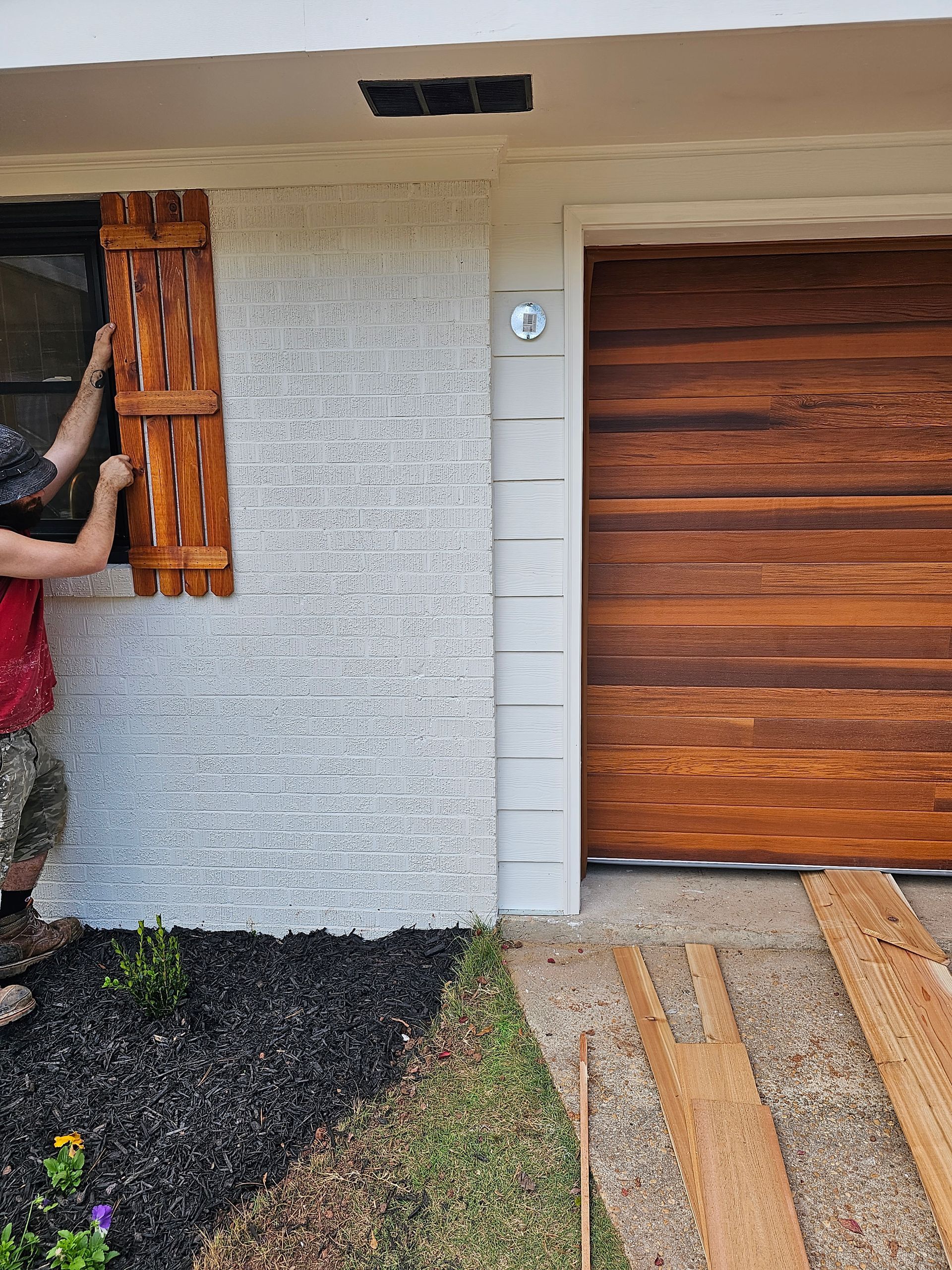 Person installing wooden shutter on a textured white exterior wall near a brown garage door.