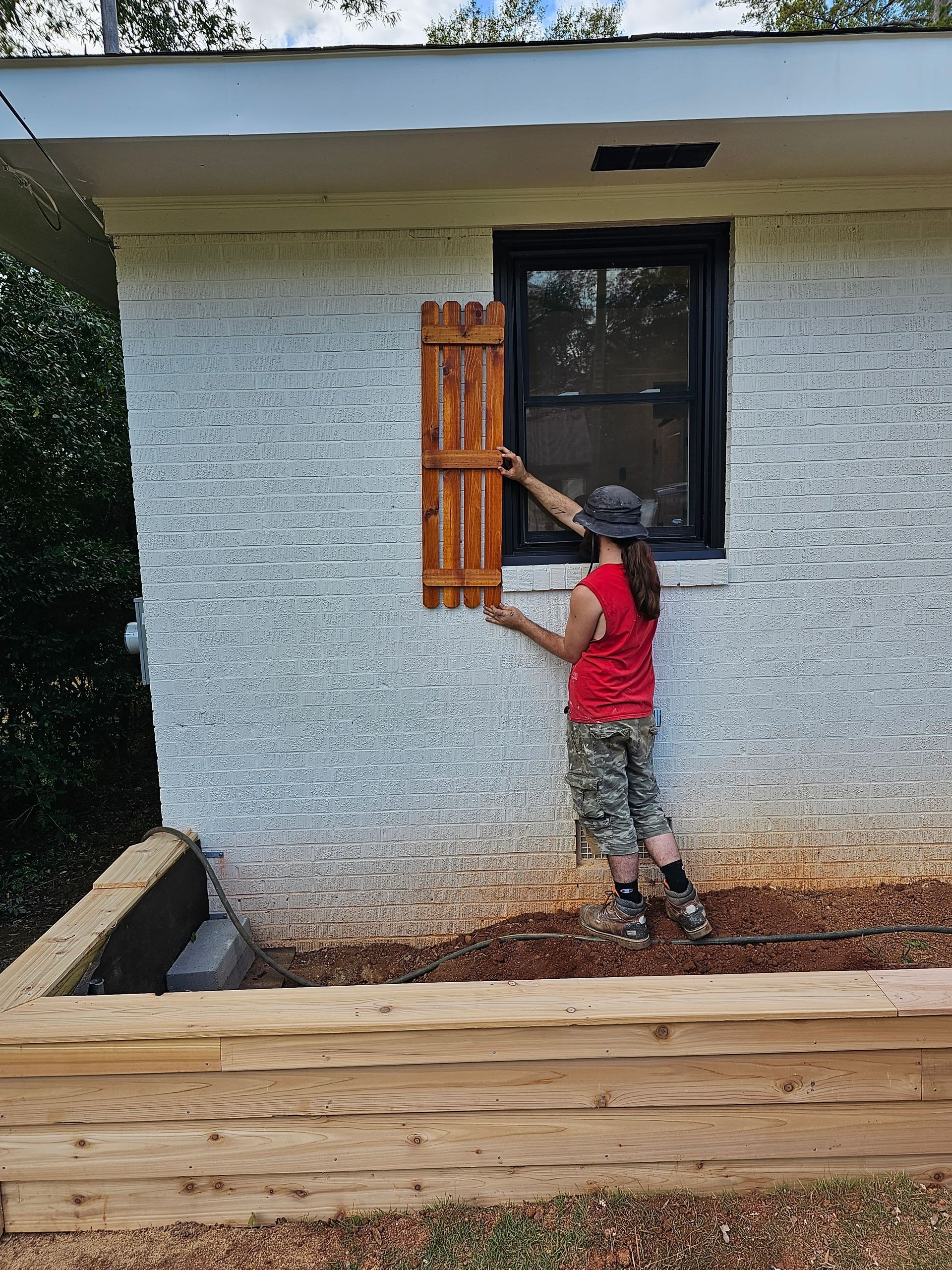 Person attaching a wooden shutter to a black-framed window on a white-painted brick wall.