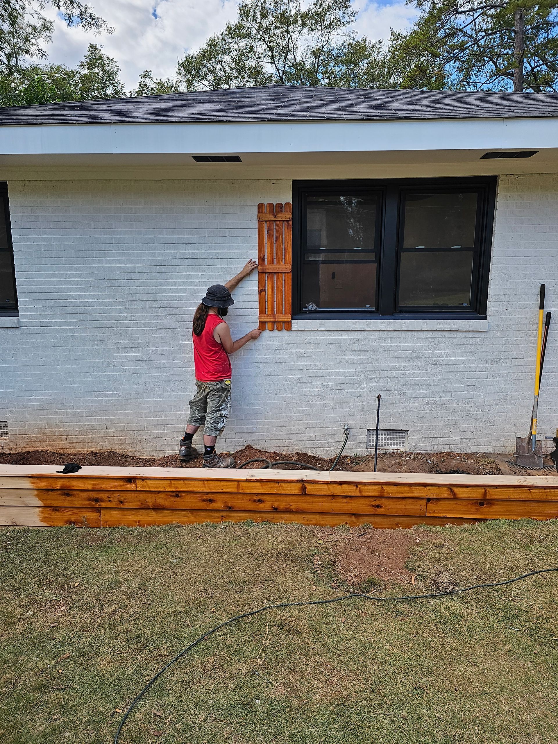 Person installing a wooden shutter on a white brick house with black windows. Brown wooden flower bed in the foreground.