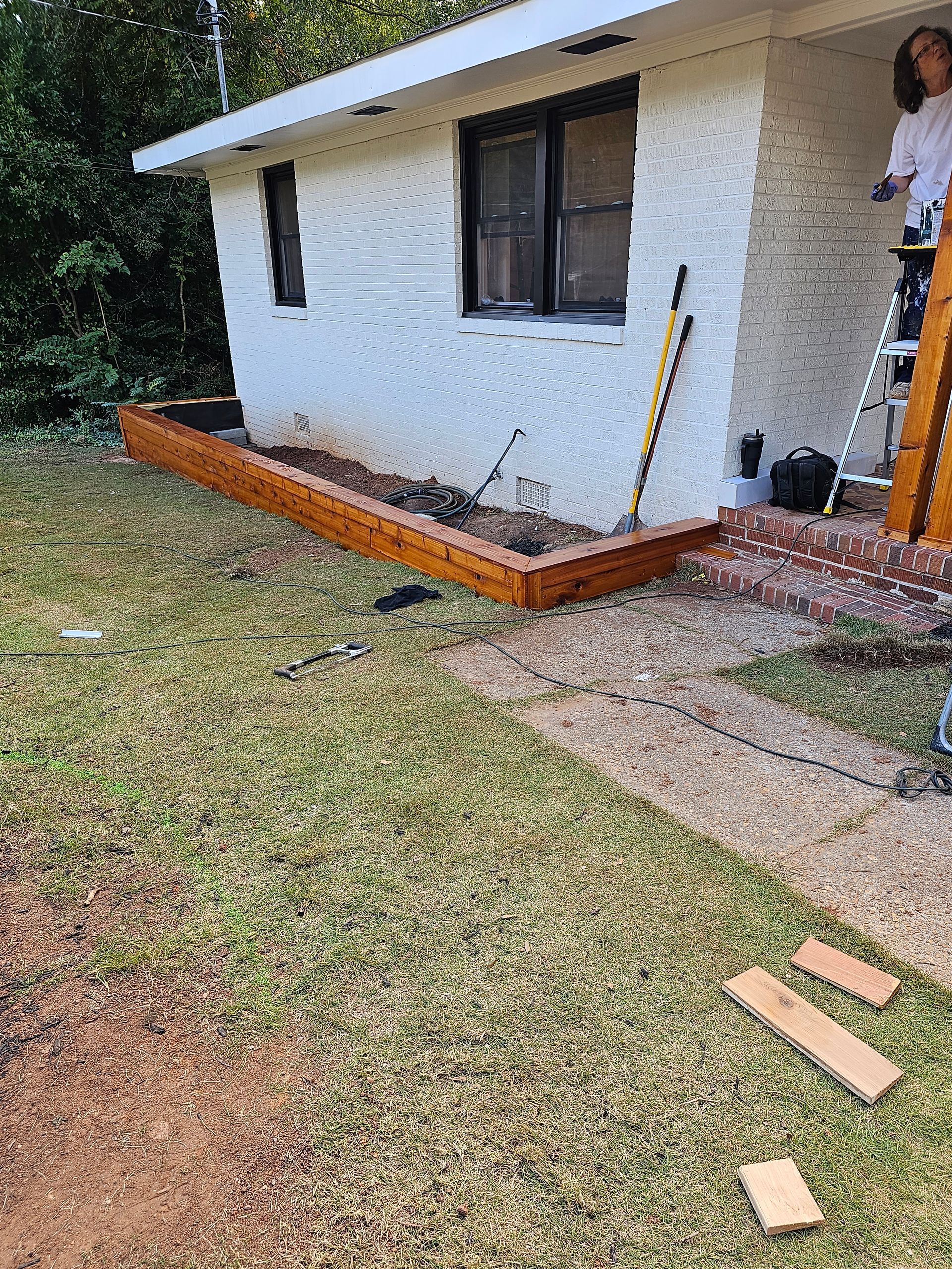 A person is on a ladder by a house. A newly built, stained wood planter box is in front.